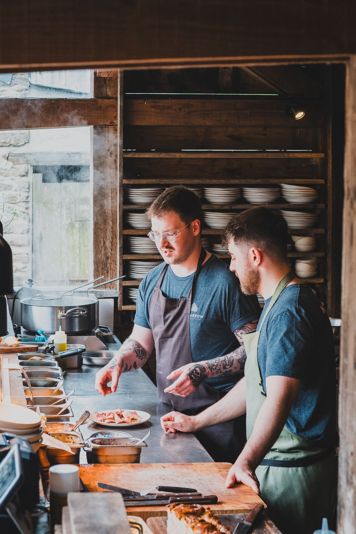 Two tattooed chefs in aprons preparing dishes at an open kitchen counter in a rustic restaurant.