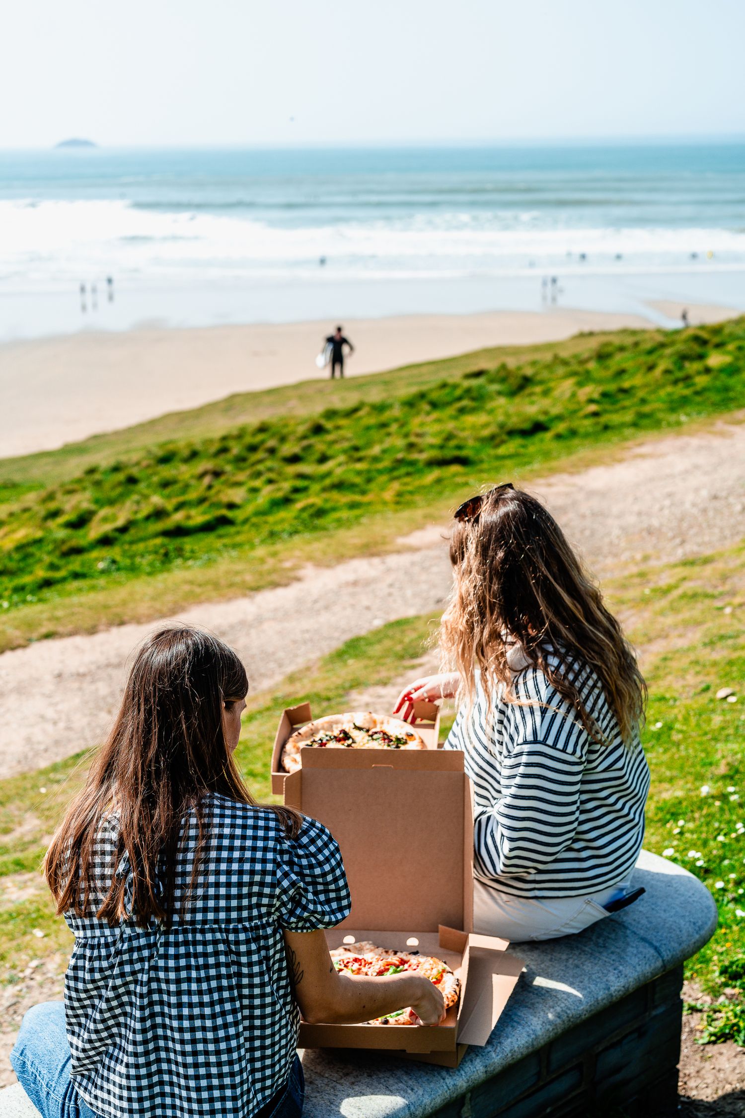 Two women enjoying pizza from takeaway boxes overlooking a sunny beach and ocean waves.