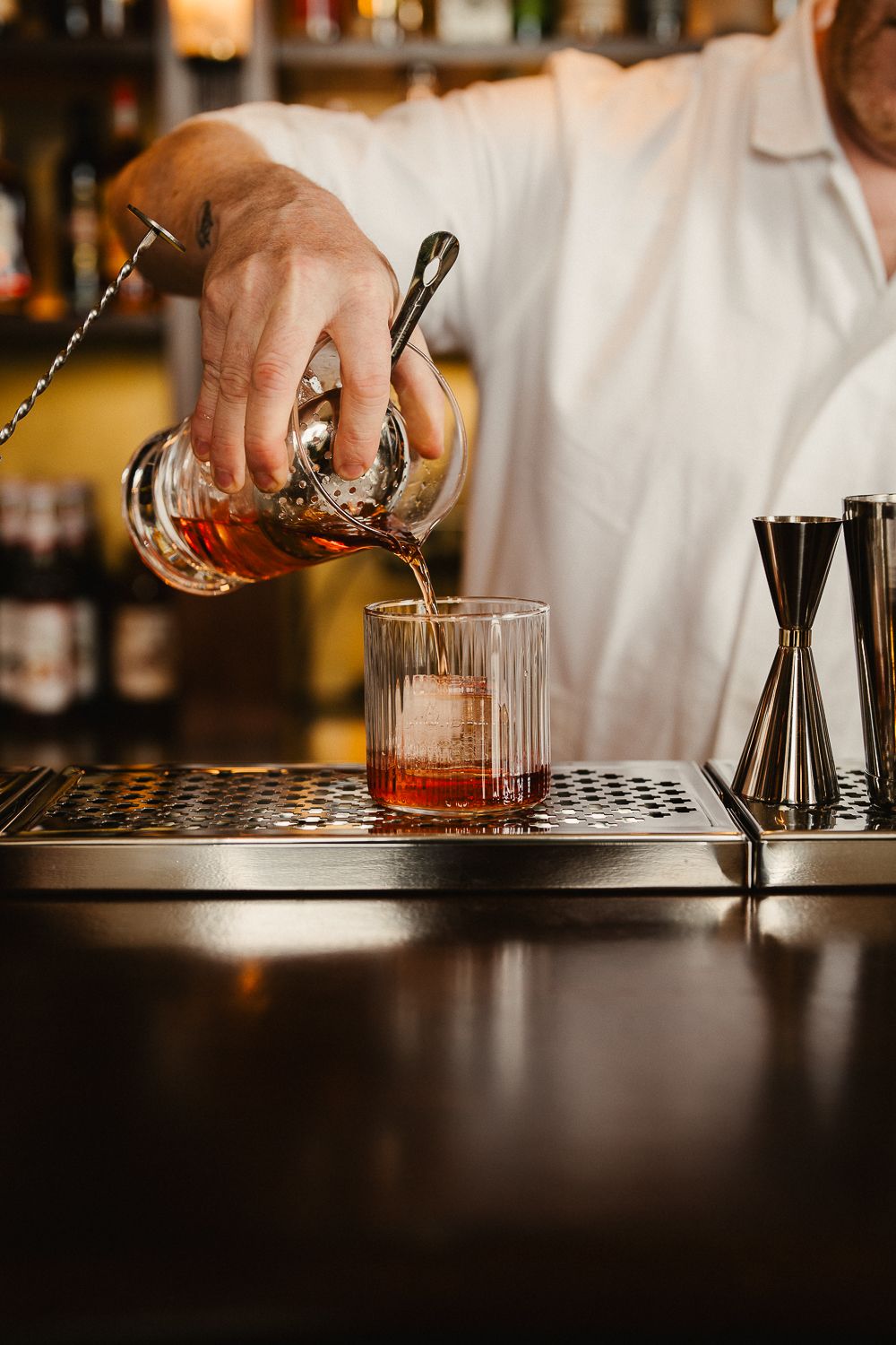 Bartender straining a cocktail over ice into a crystal glass at a bar.