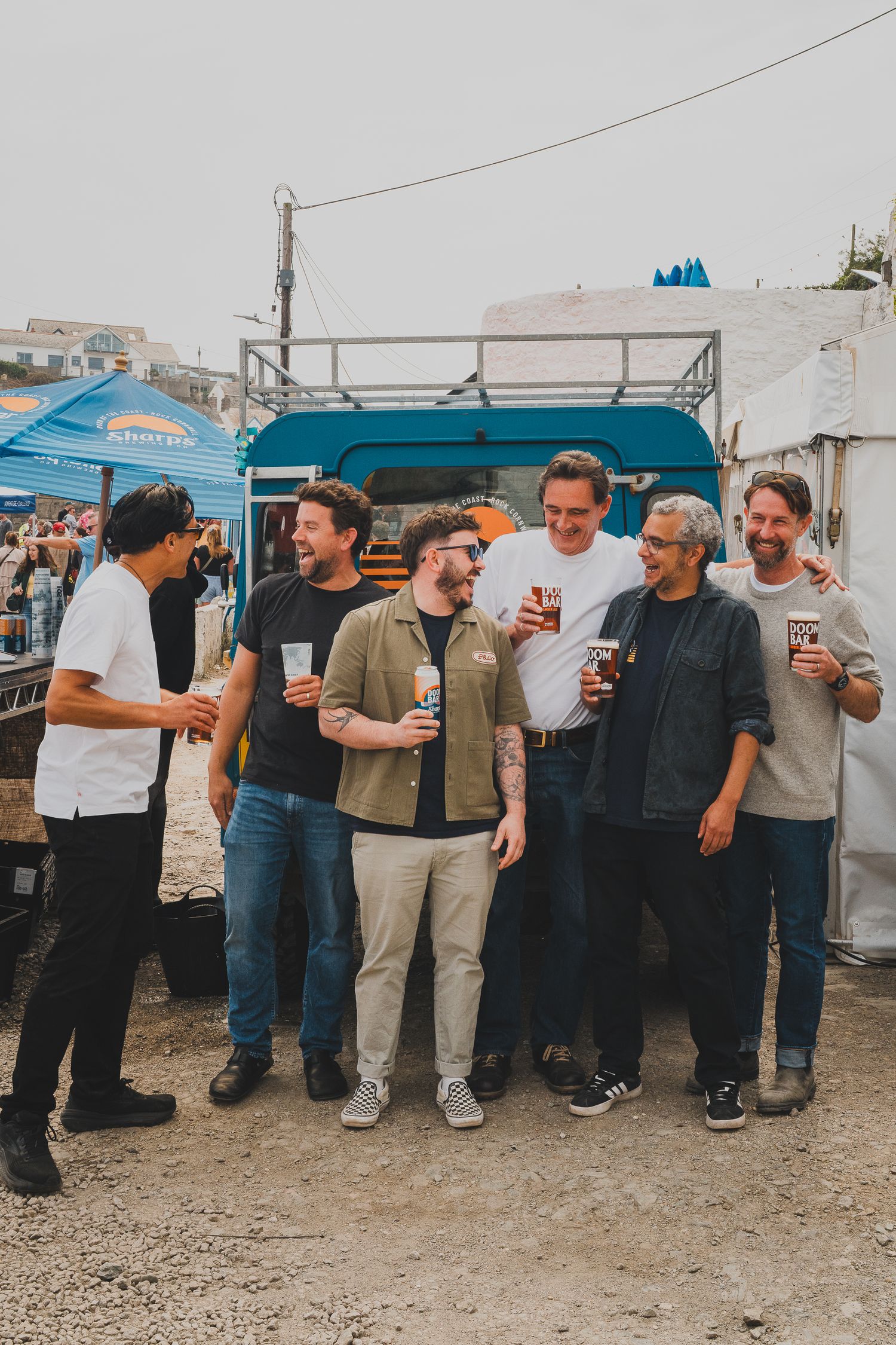 Group of six men laughing and holding drinks at an outdoor food festival near a blue truck.