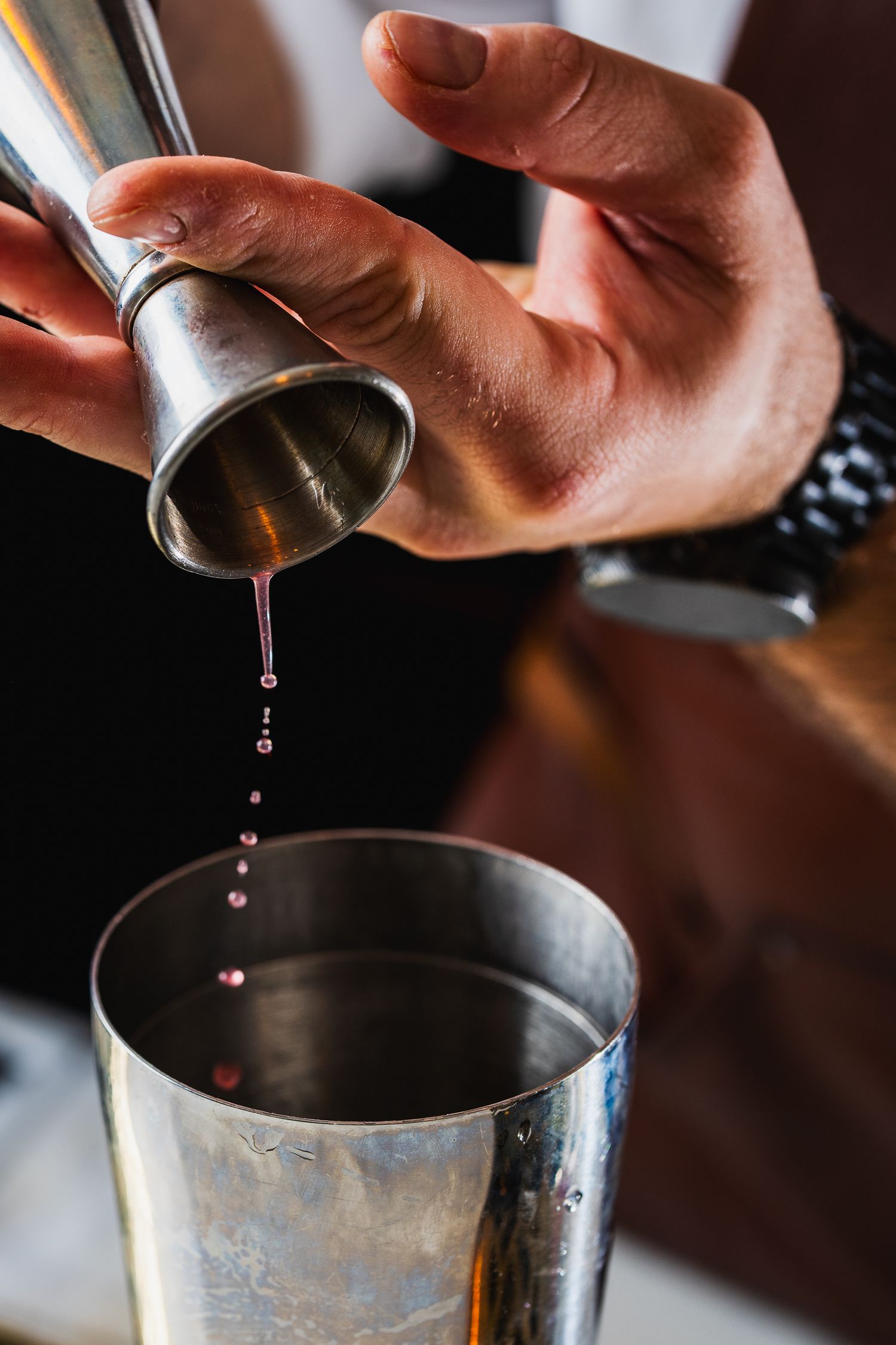 Bartender pouring pink liquid from jigger into metal cocktail shaker for drink preparation.