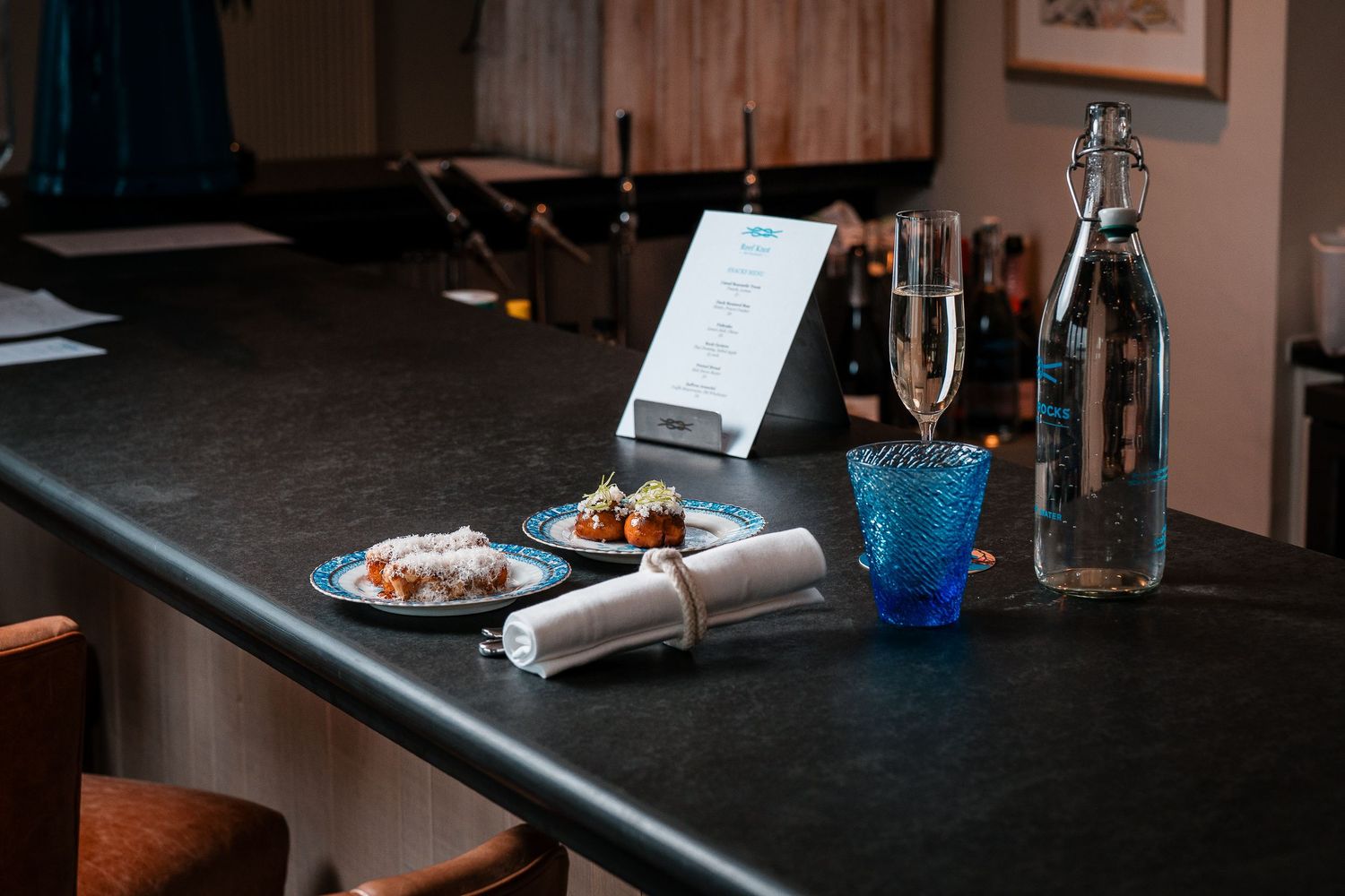 Elegant bar counter setting with blue patterned plates, champagne flute, blue glass, and water bottle with menu stand.