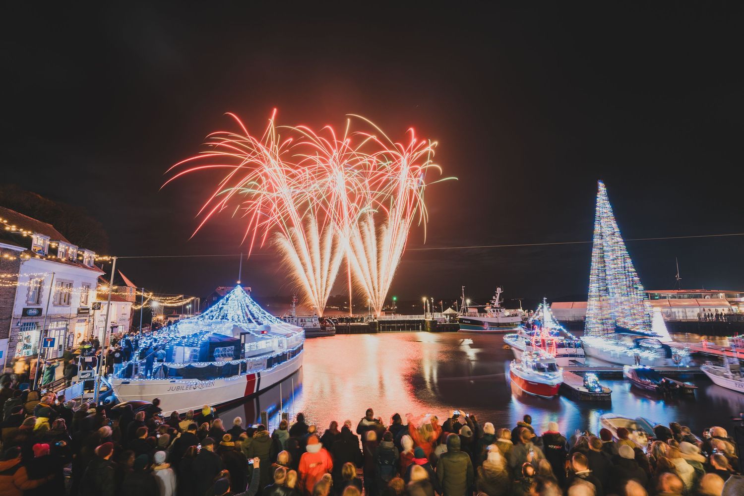 Spectacular fireworks display over a harbor with Christmas trees and crowds at night.
