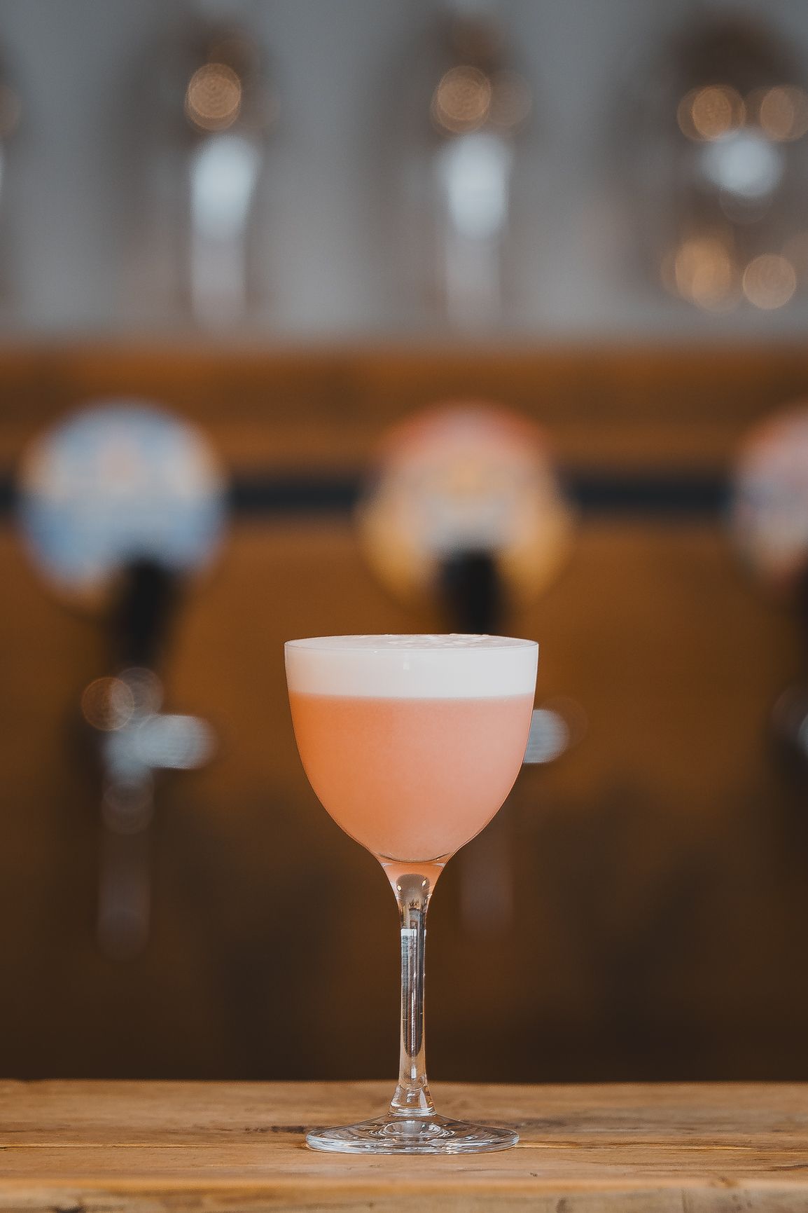 Elegant pink cocktail with frothy foam top in coupe glass on wooden bar with tap handles background.