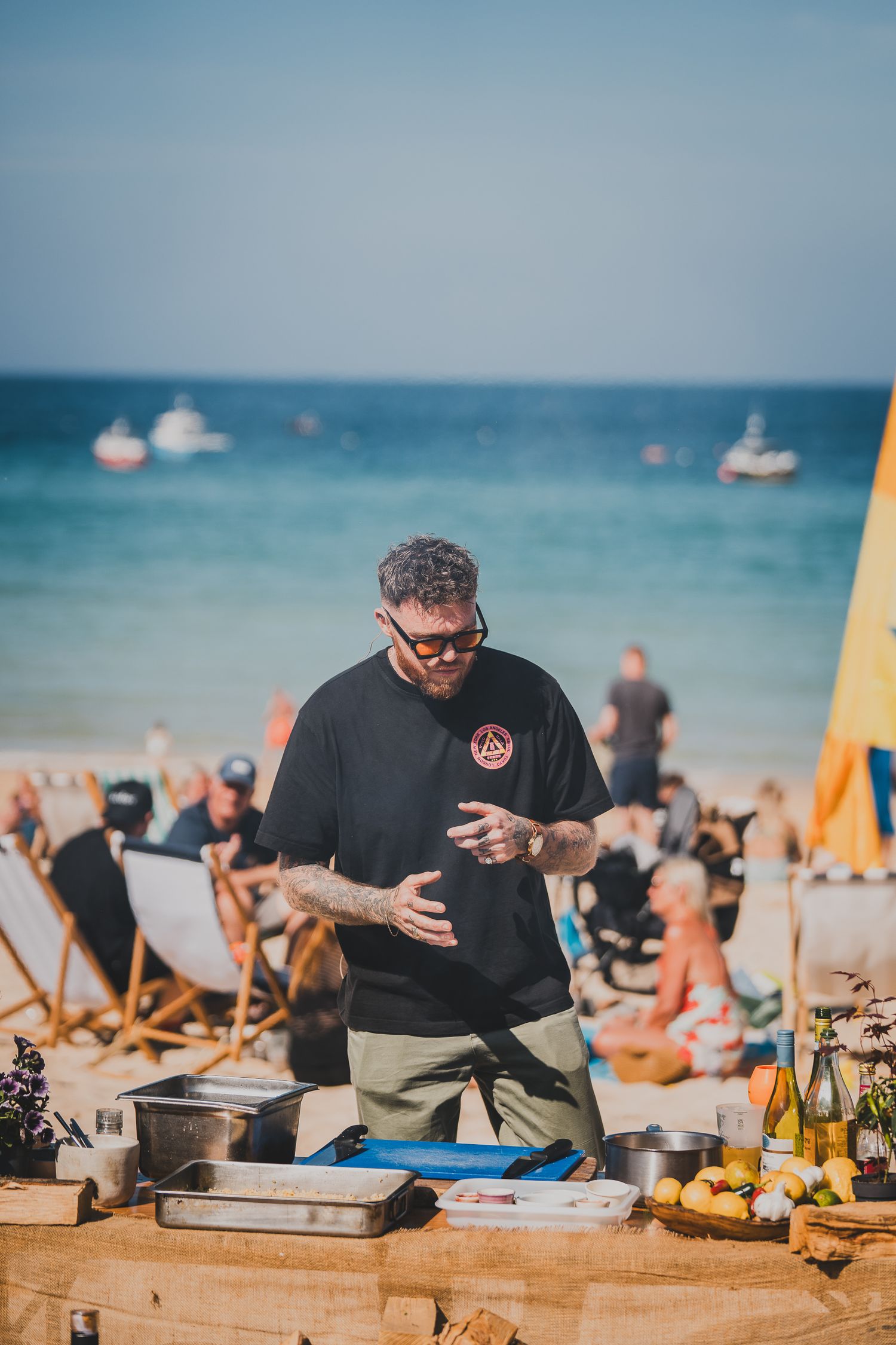 Tattooed chef in black shirt prepares food at a beachside cooking station on a sunny day.