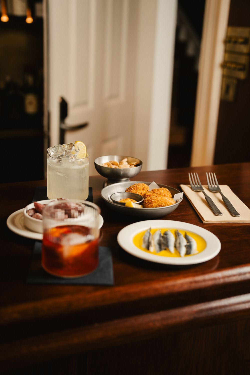 Bar spread of cocktails, anchovies, croquetas and octopus tapas on dark wooden counter.