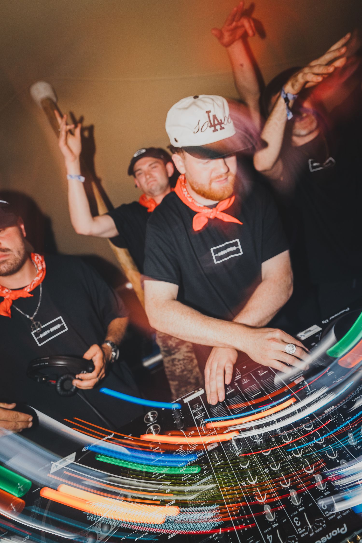 A bearded DJ in an LA cap works Pioneer decks with motion blur effects and crowd behind him.