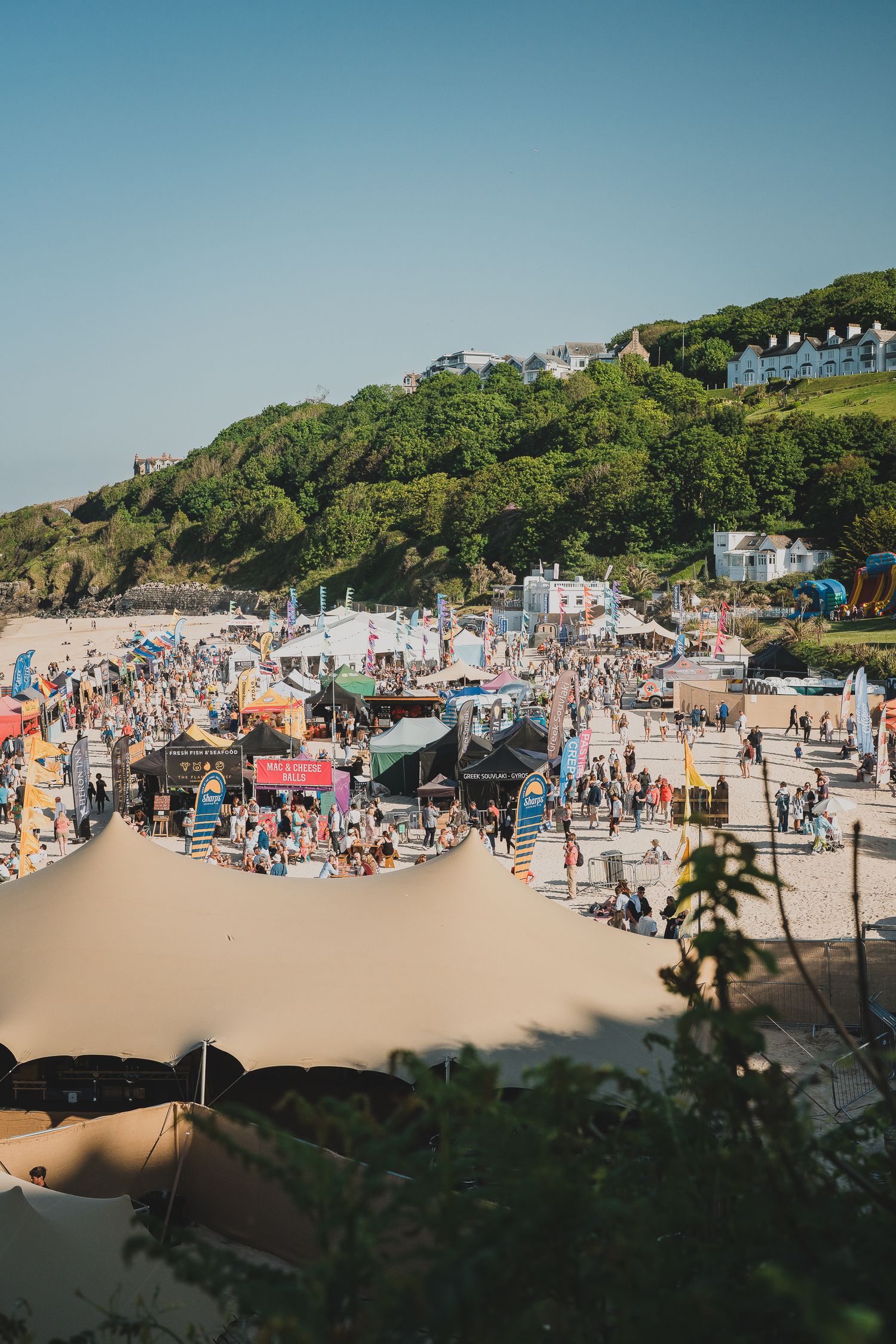 Aerial view of busy beach food festival with vendor stalls, crowds, and green hillside backdrop.