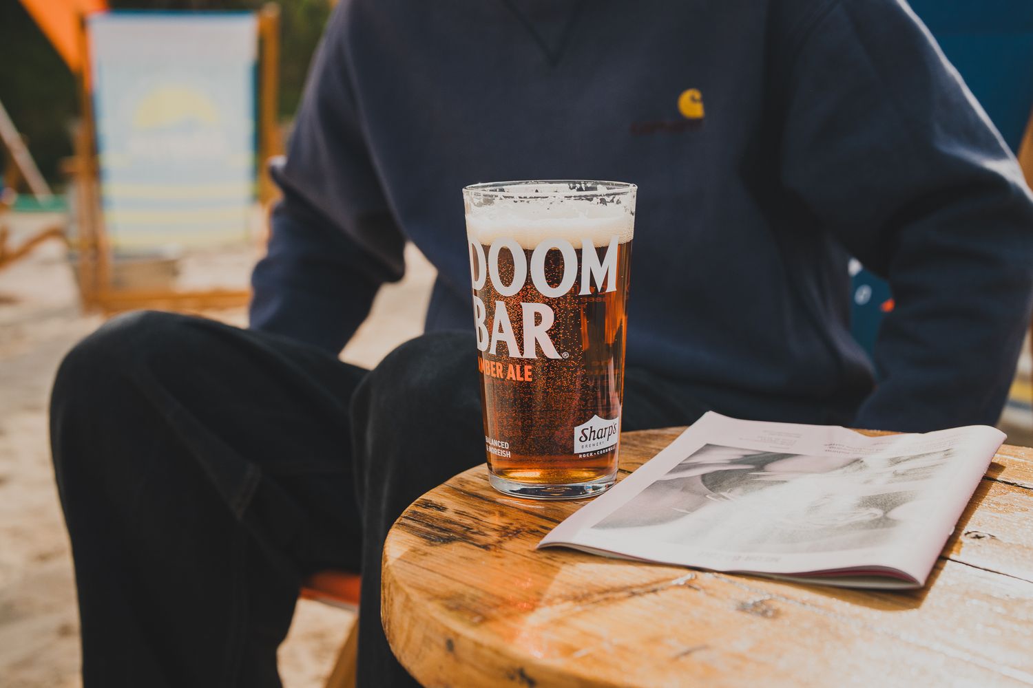 Person holding a pint of Doom Bar real ale at an outdoor wooden table with magazine.