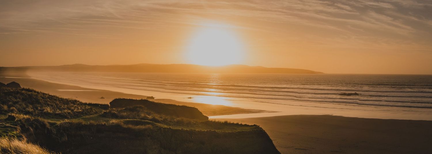 Panoramic golden sunset over a coastal beach landscape with rolling hills and calm ocean waves.