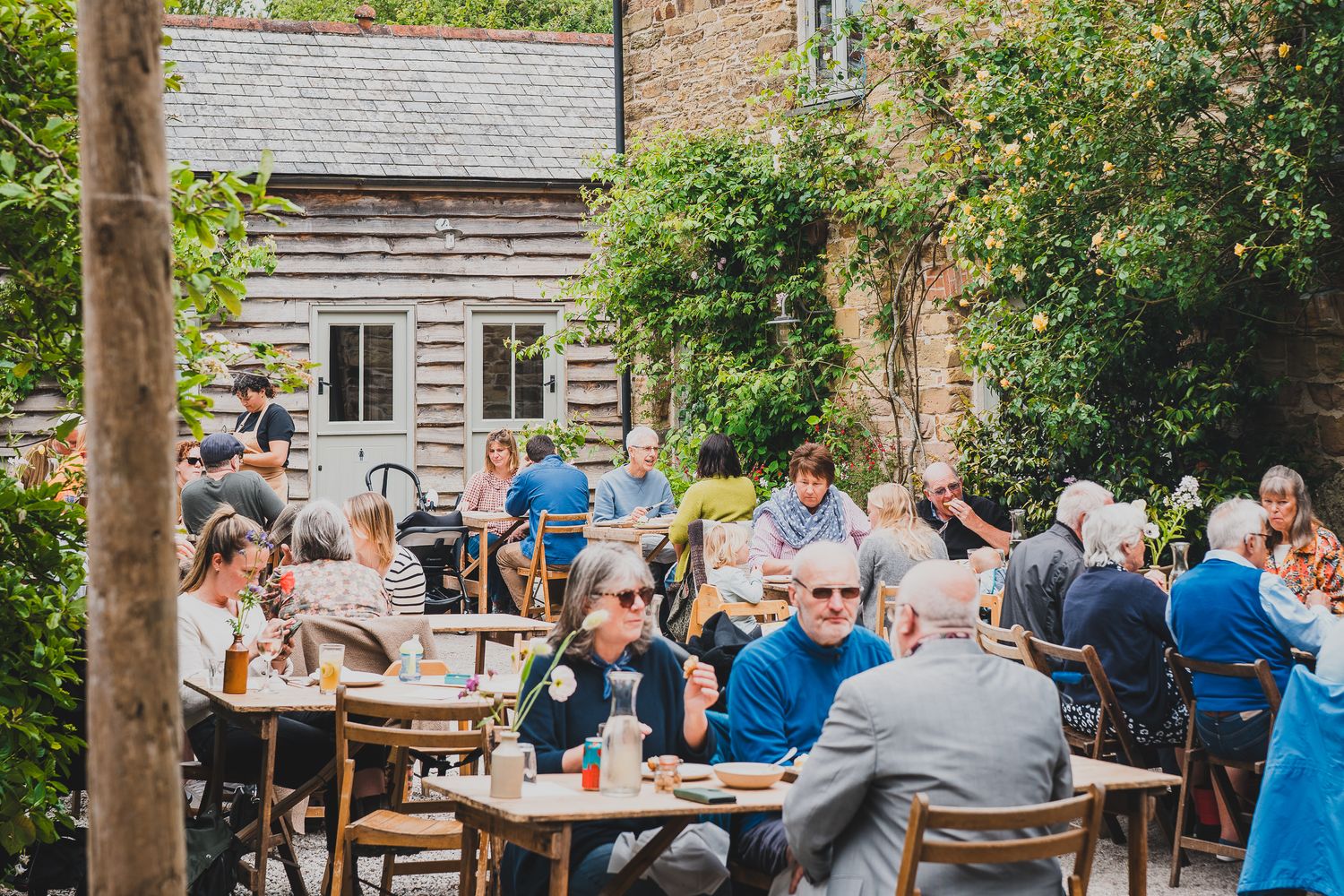 Busy outdoor restaurant patio with diners seated at wooden tables surrounded by trees and greenery.