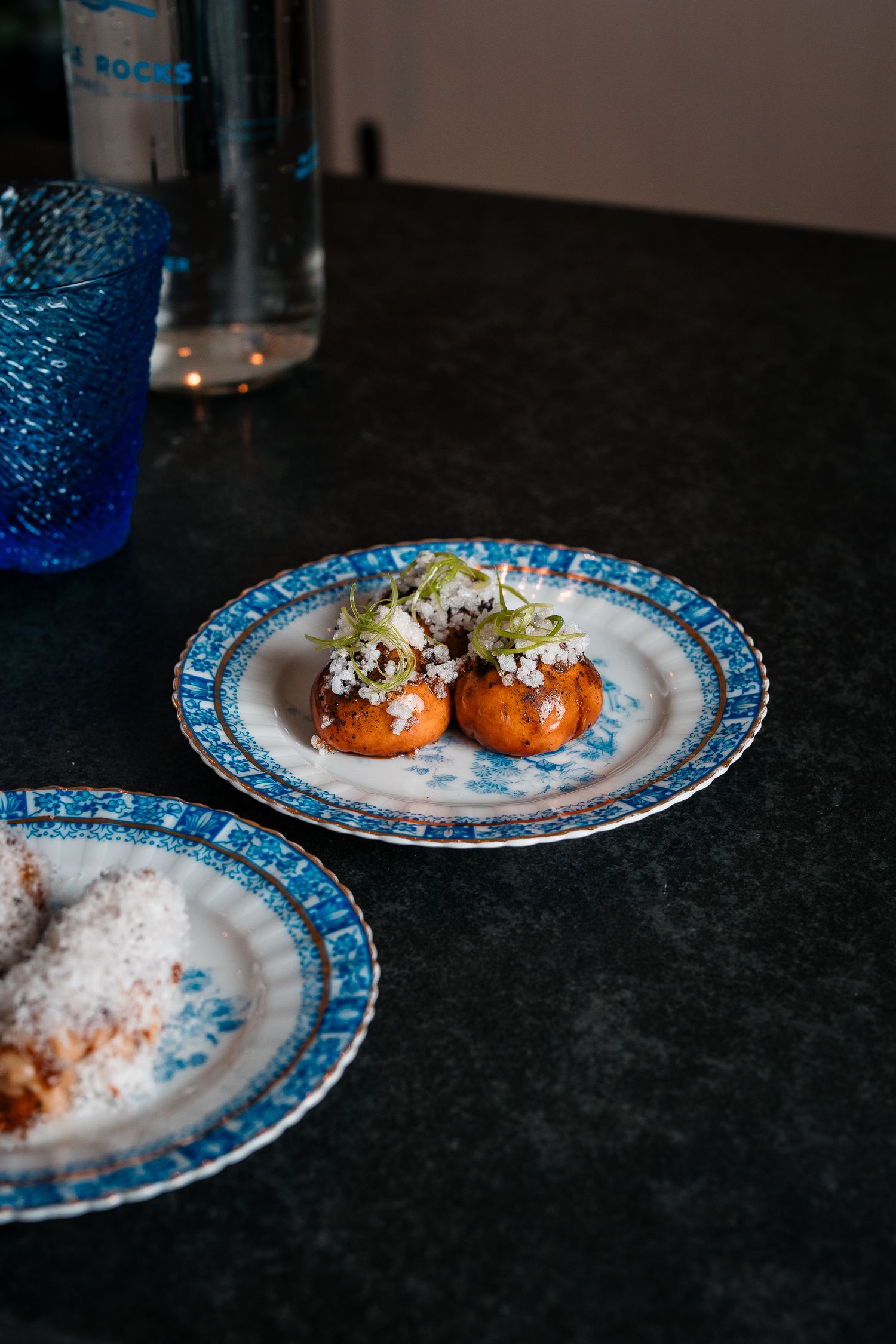 Blue and white decorative plates with golden fried arancini topped with crumbled cheese and microgreens on dark counter.