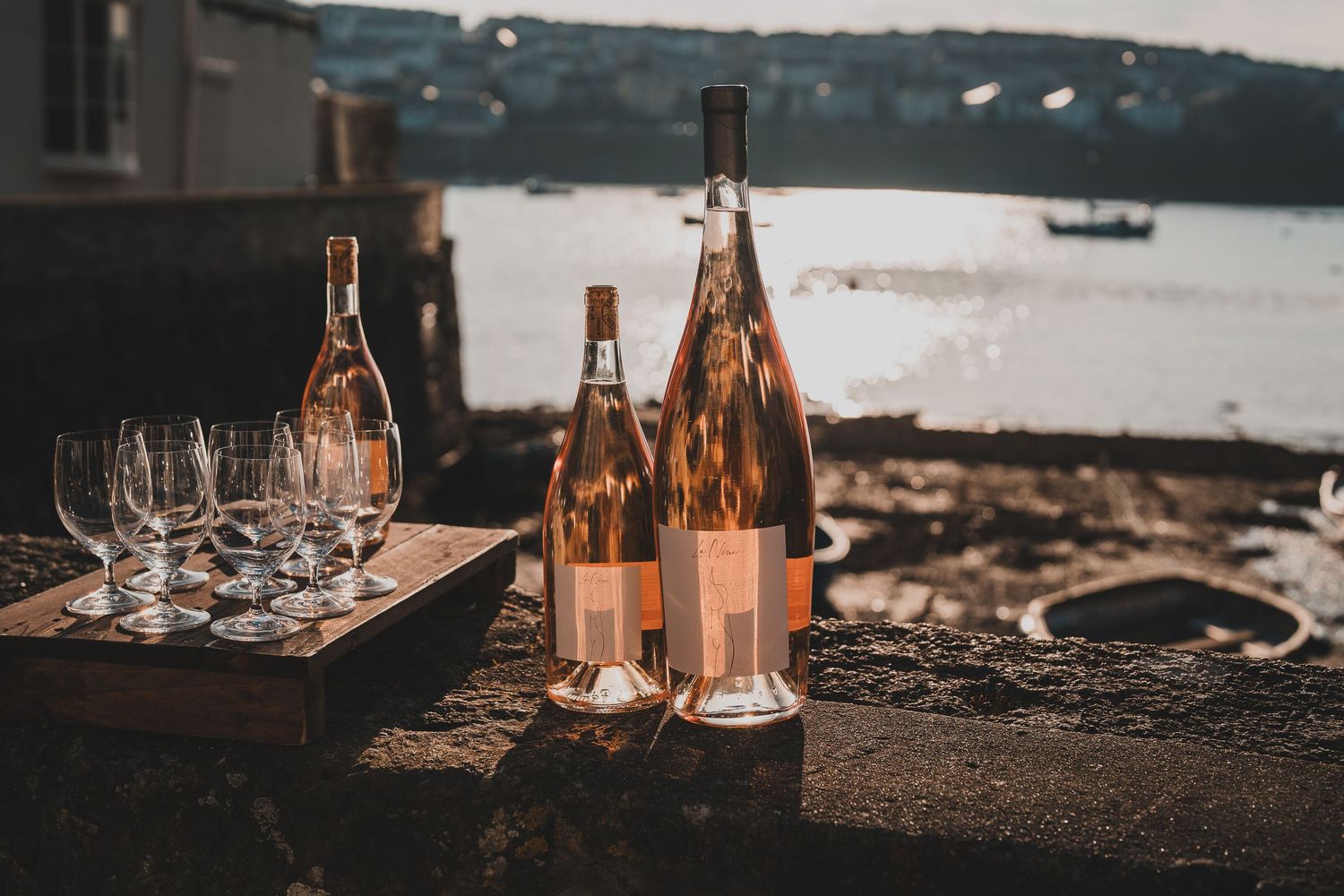 Rose wine bottles and glasses arranged on a stone wall with a scenic harbor view at dusk.