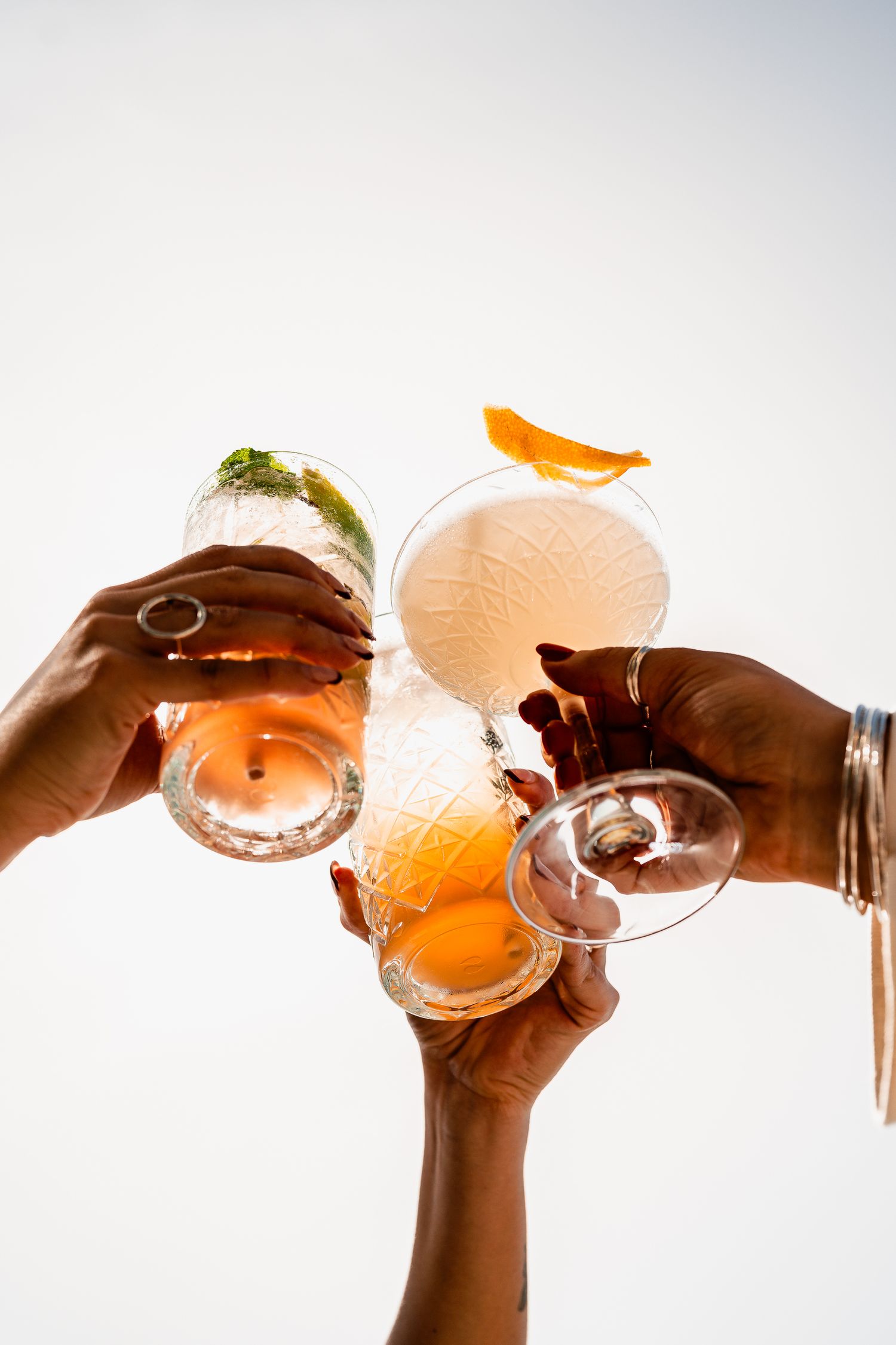 Three hands toasting with colorful citrus cocktails against bright white background.