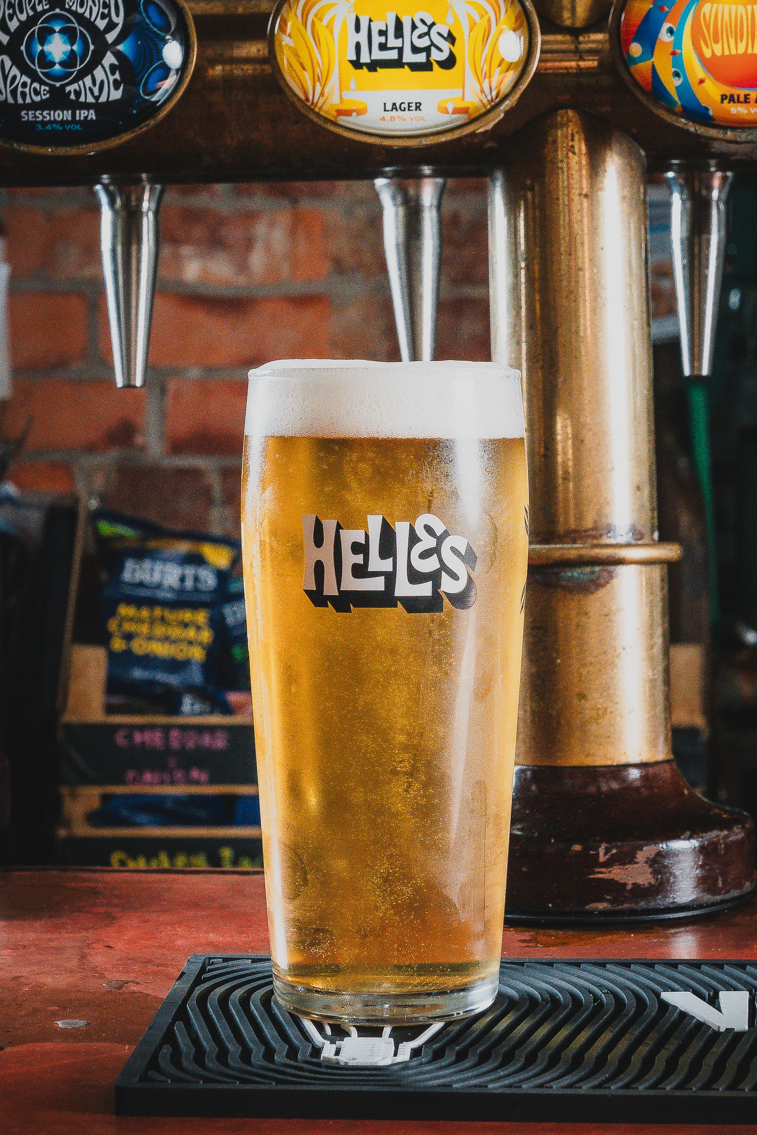 Freshly poured Helles lager in branded pint glass on bar mat with brick wall background.
