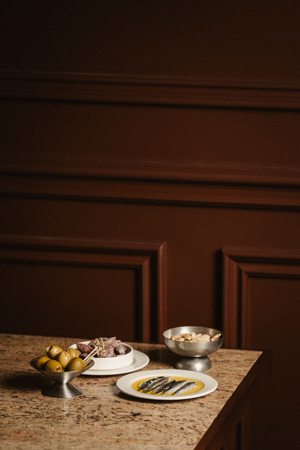 Elegant marble counter with tapas dishes including olives, anchovies, and charcuterie against dark wood paneling.