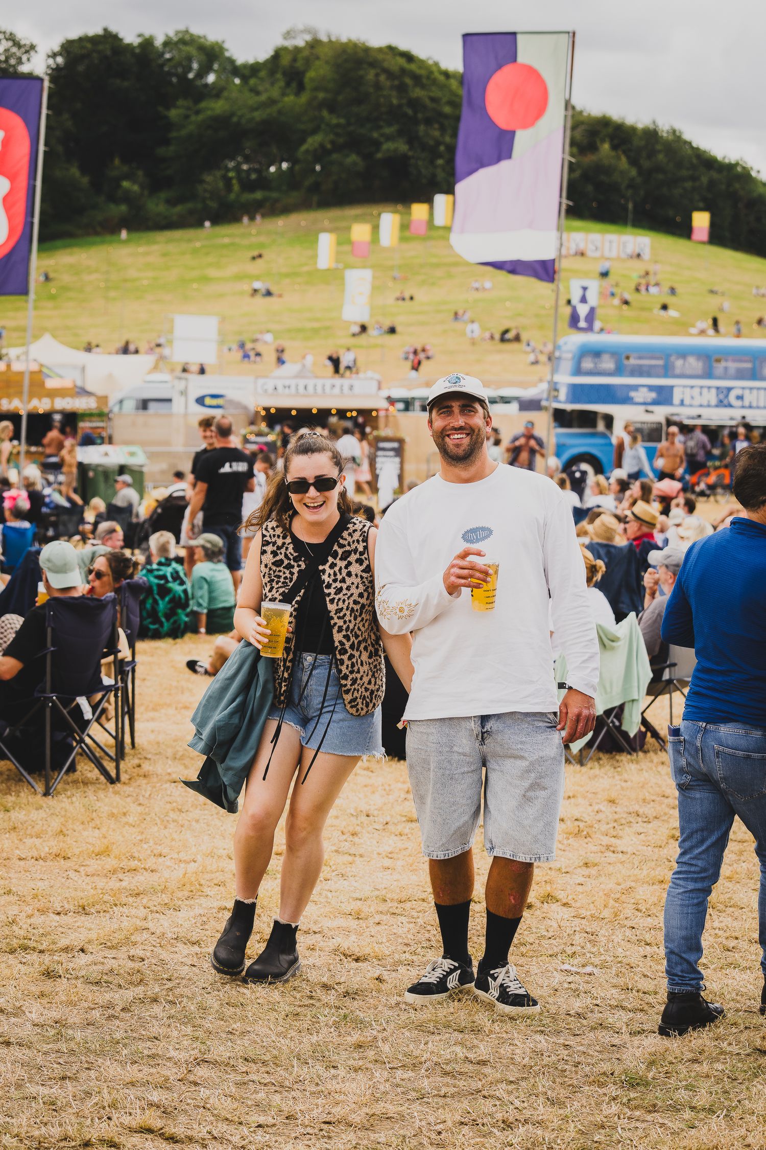 A smiling couple holding beer cups walks through a sunny outdoor music festival with colorful flags.