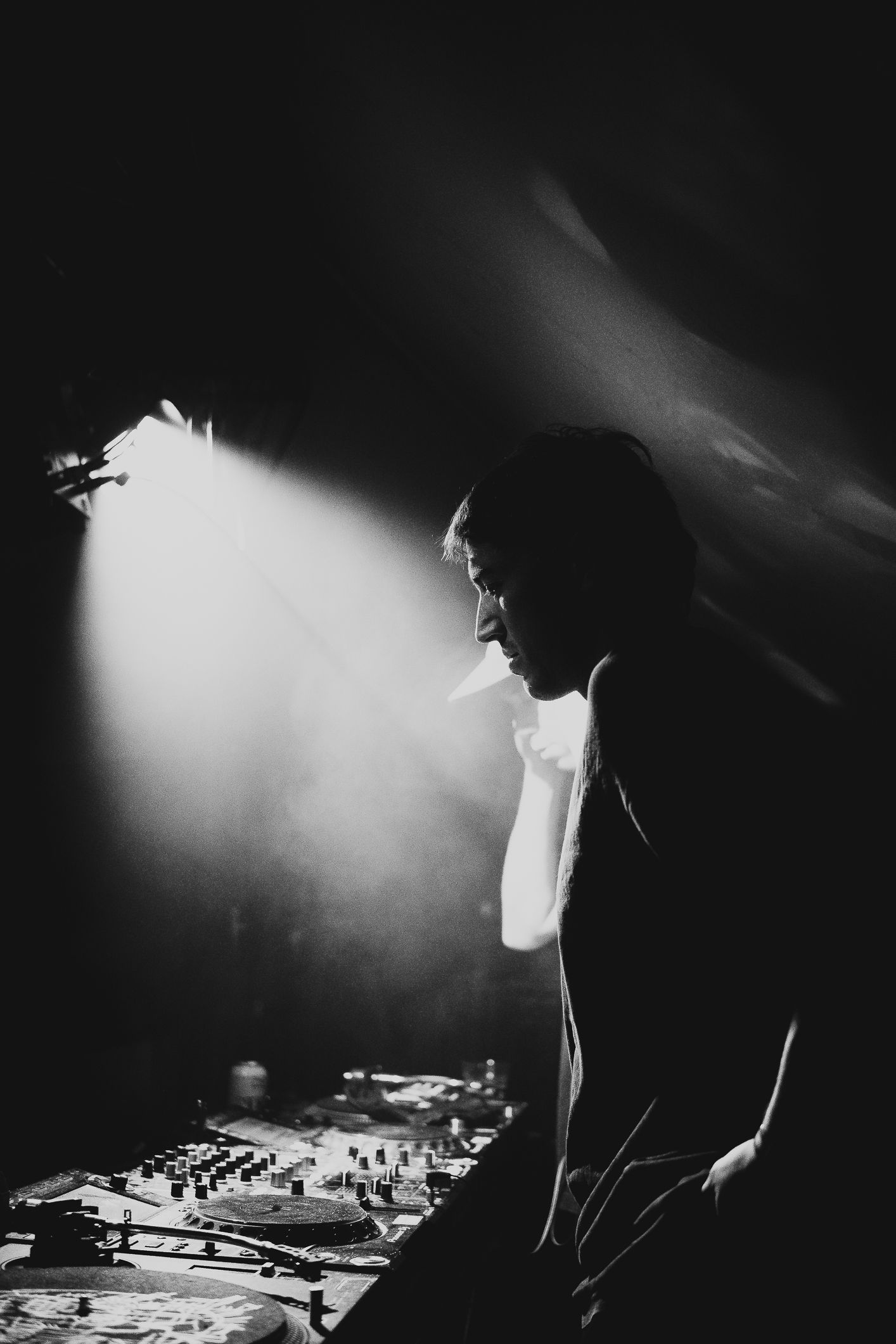 Black and white silhouette of a DJ performing at turntables under a dramatic spotlight beam.