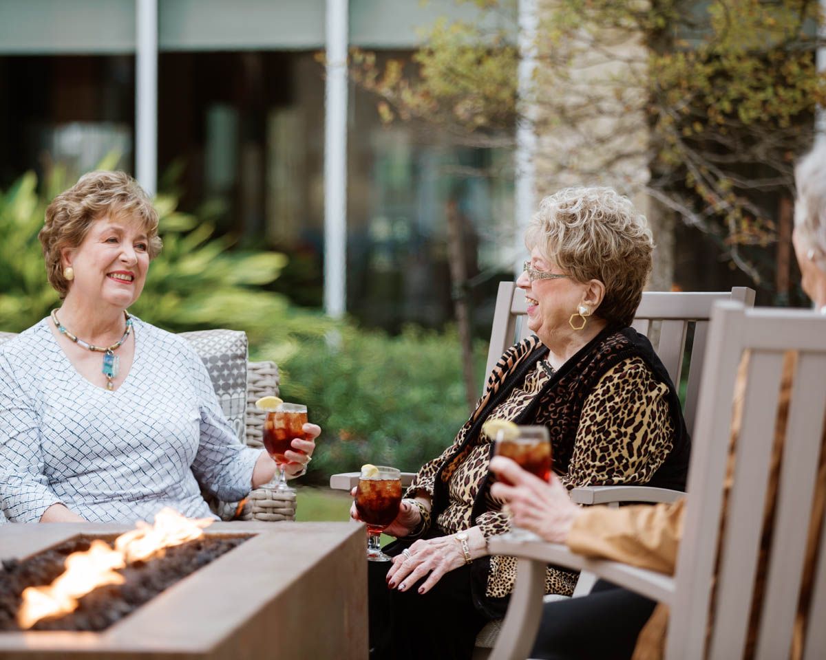Three elderly women socializing outdoors around a fire pit, enjoying drinks and conversation at a senior living community.