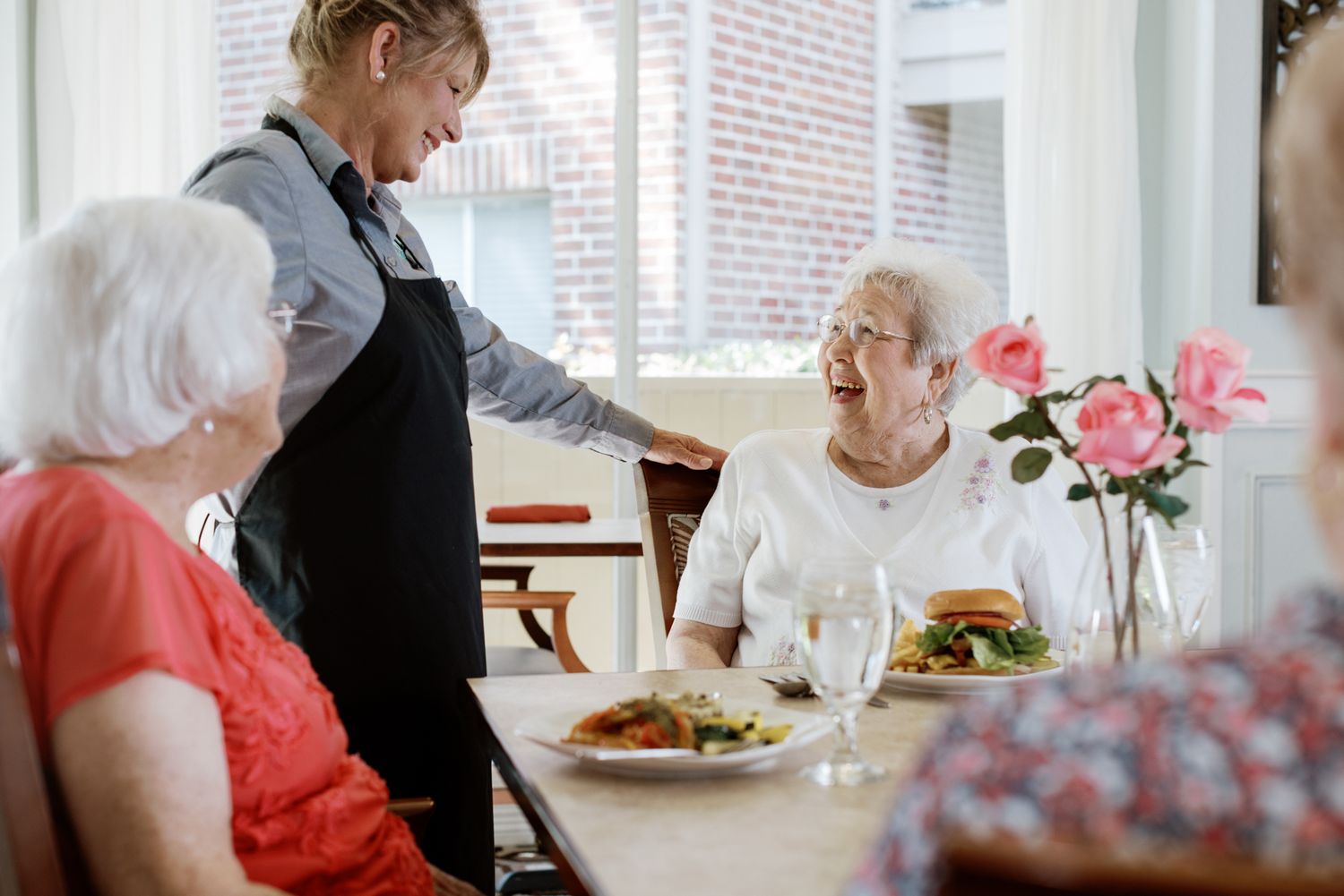 A caregiver serving meals to happy elderly women at a dining table with fresh flowers in a bright room.