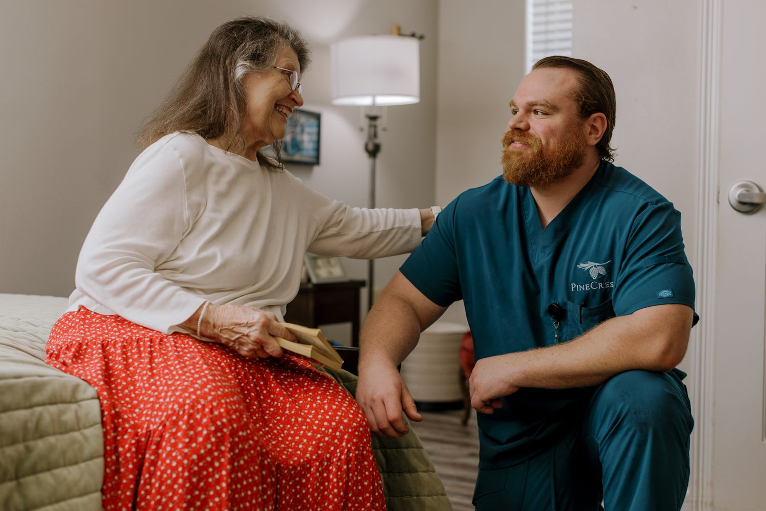 Healthcare worker in scrubs kneeling beside elderly woman sitting on bed during in-home medical consultation.