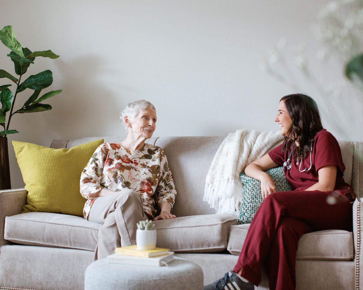Elderly woman and female caregiver in scrubs sitting together on a couch, smiling and conversing in a bright living room.