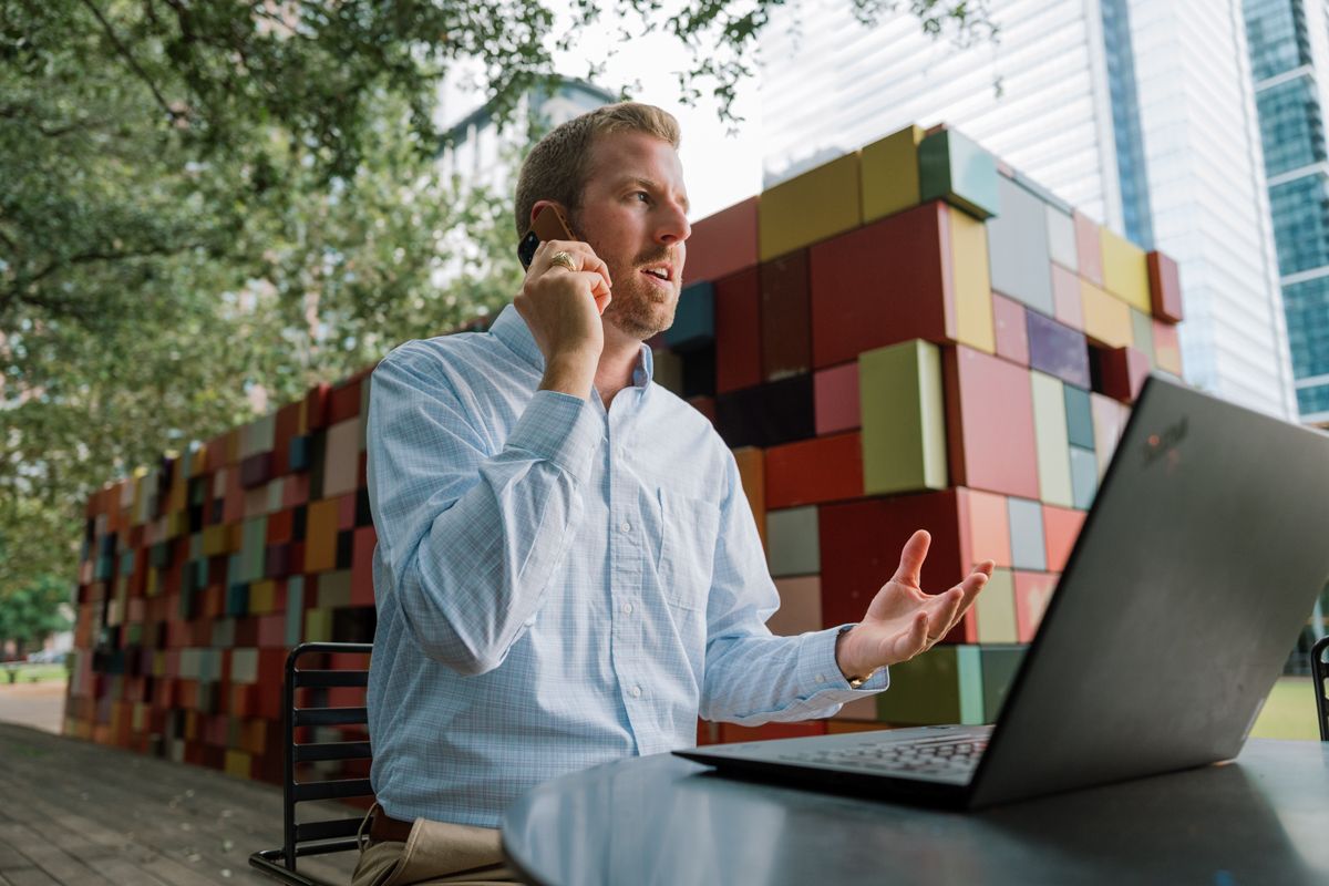 Young professional talking on phone while working on laptop outdoors near colorful building.