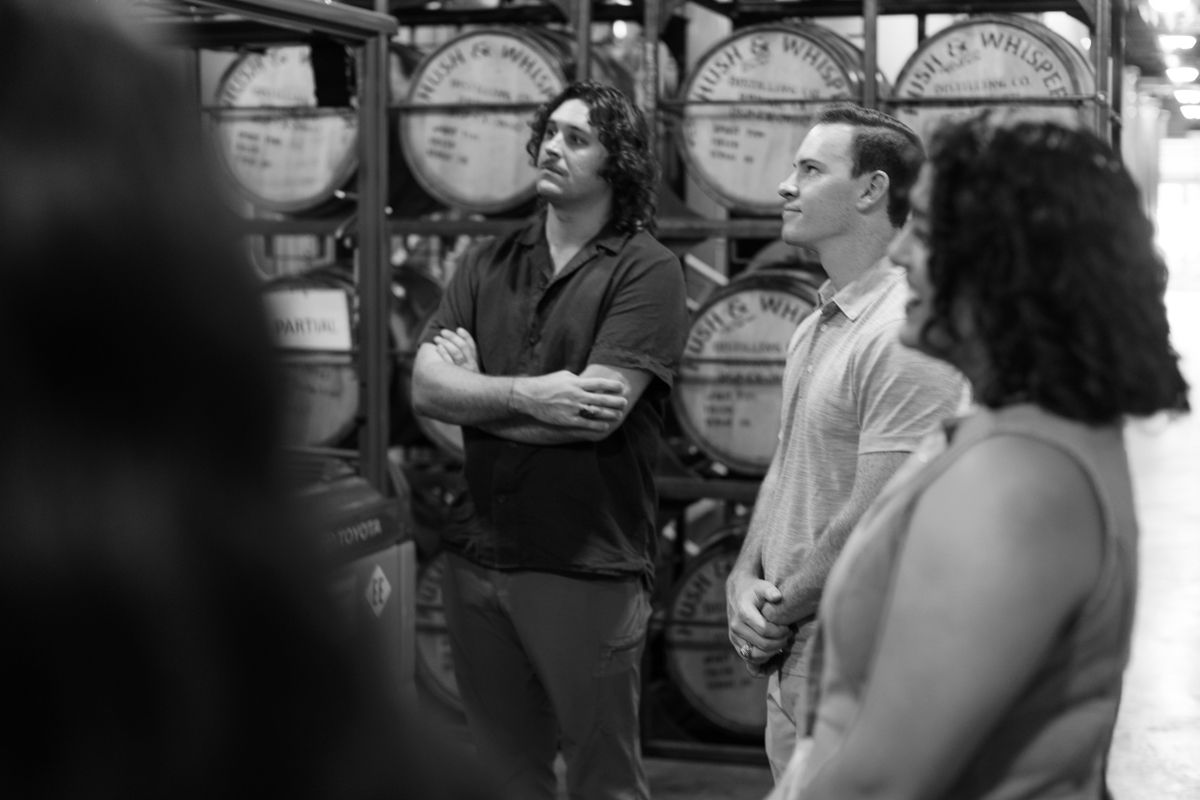 Black and white photo of three people conversing near whiskey barrels in a distillery warehouse.