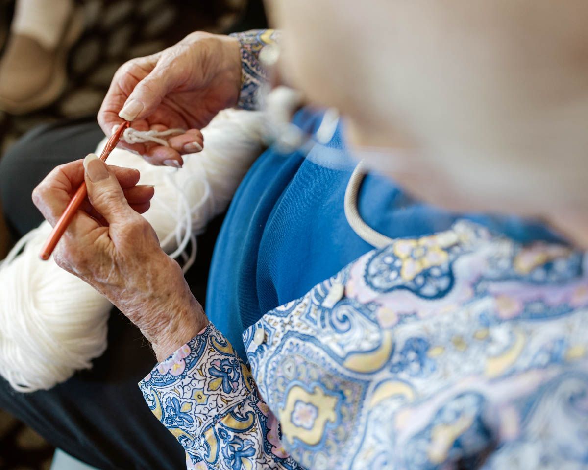 Close-up of elderly woman's hands knitting with orange needles and white yarn in a floral blouse.