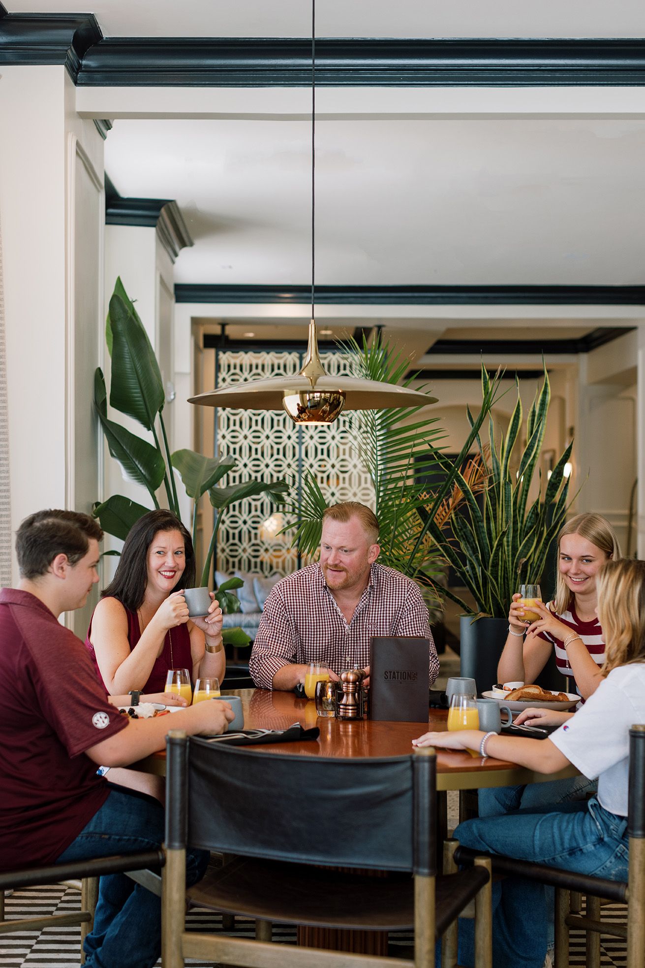 Group of friends laughing and dining together at a stylish restaurant with tropical plants and warm lighting.