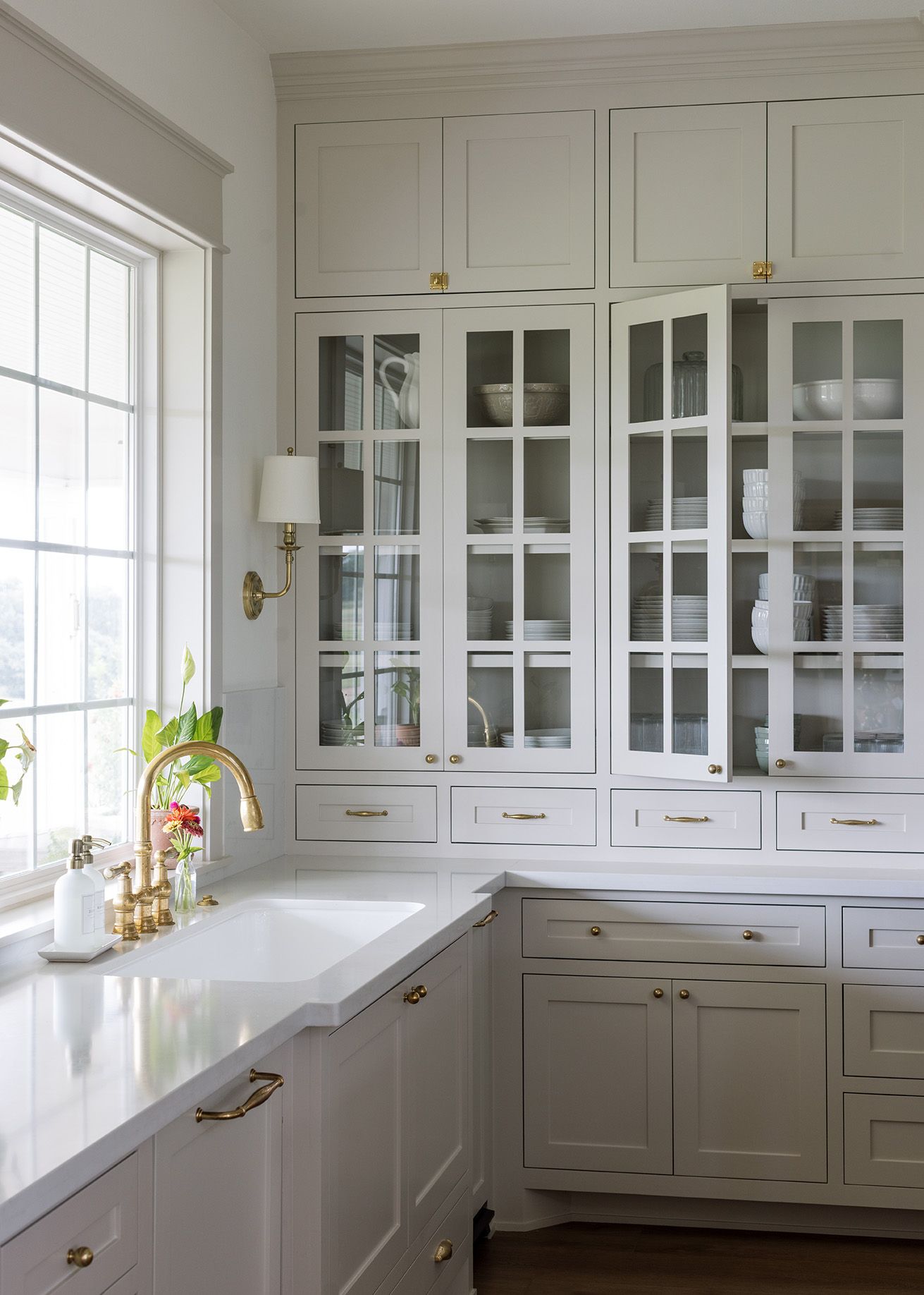 Floor-to-ceiling light gray kitchen cabinets with glass fronts, gold hardware, and farmhouse sink.