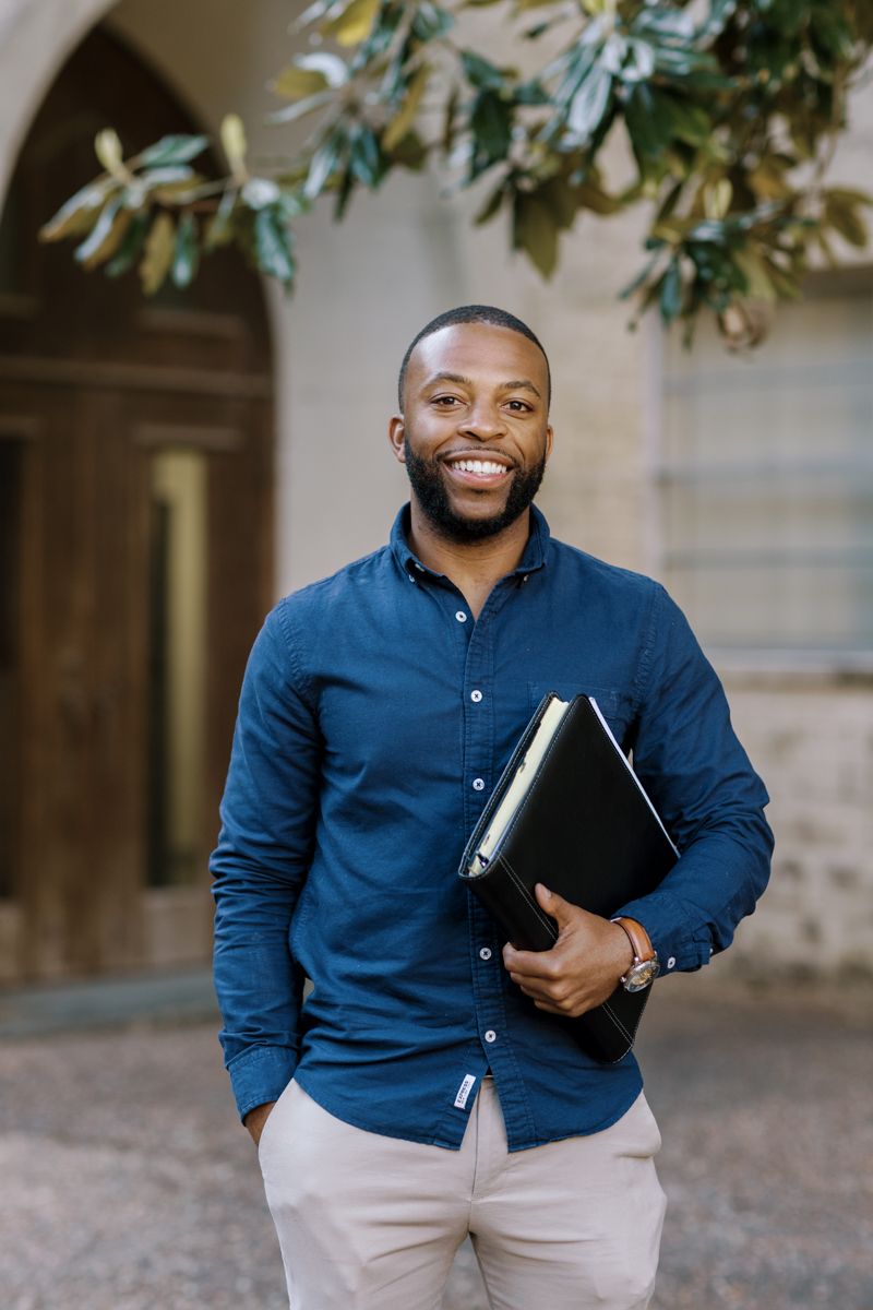 Smiling Black man in blue shirt holding a black book outside an arched stone building.