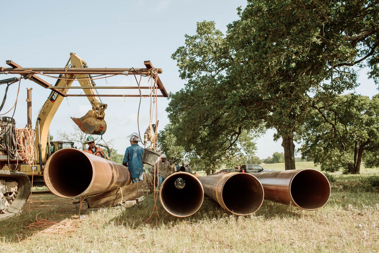 Worker stands beside large rusty industrial pipes near a yellow excavator in a grassy field with oak trees.