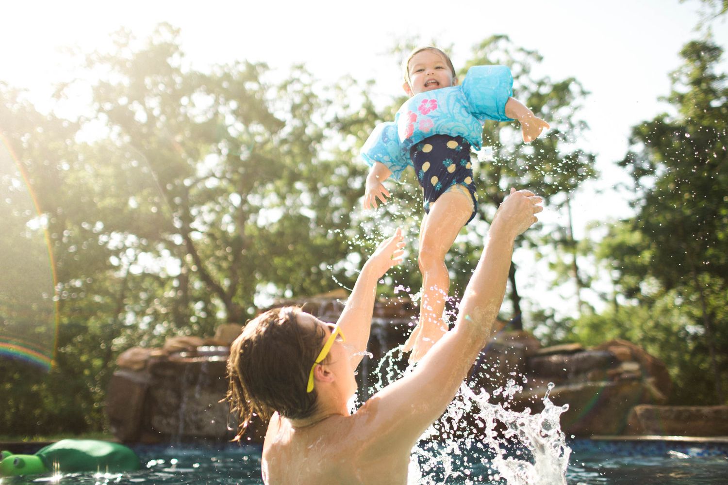 Adult tossing a happy baby in the air in a sunny outdoor swimming pool, with trees in the background.