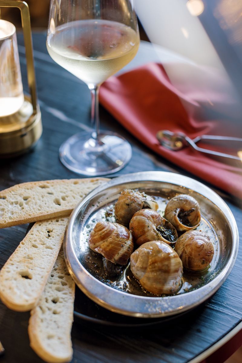 Escargot snails in butter sauce on silver plate with crusty bread slices and white wine glass on dark table.