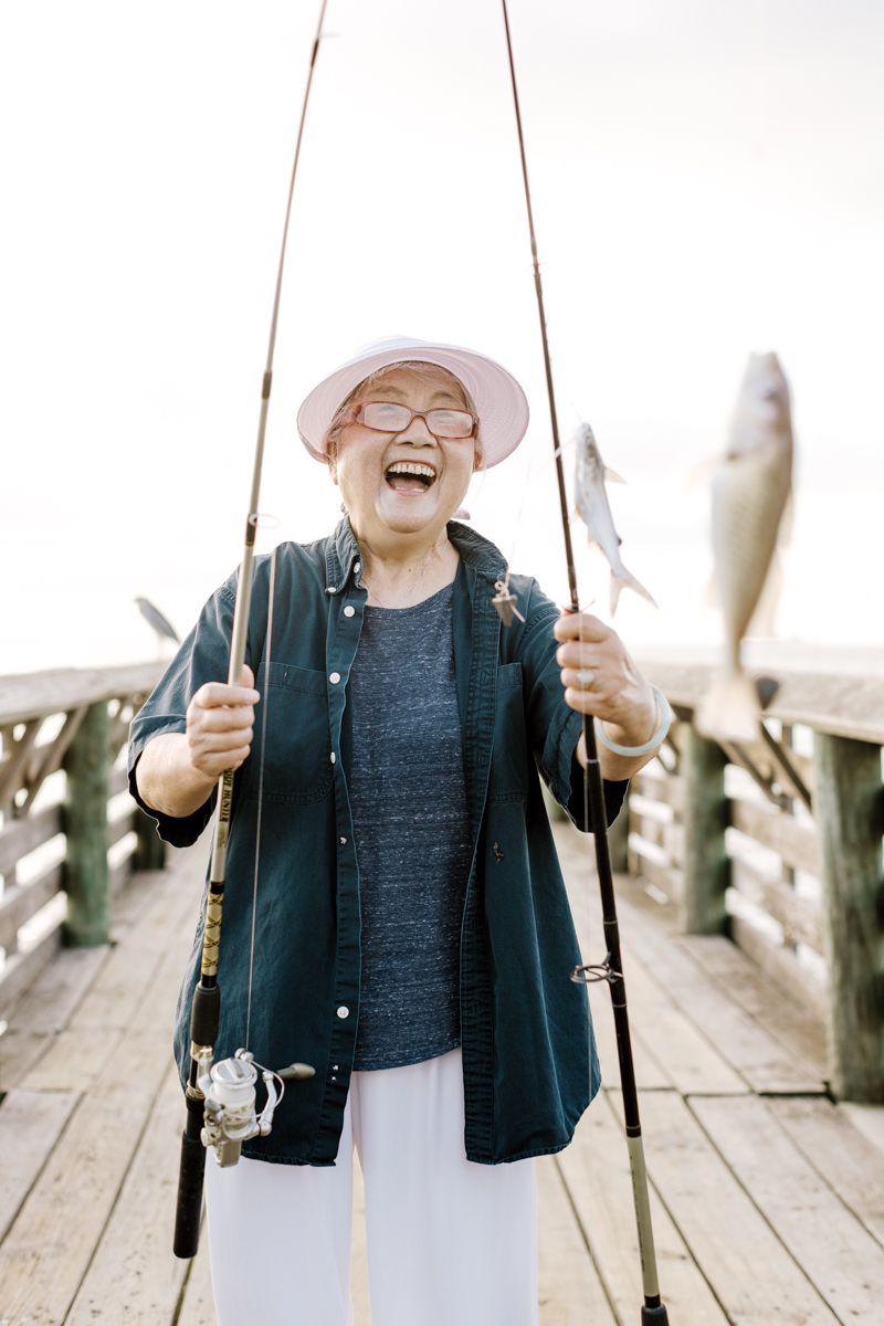 Joyful senior Asian woman laughing on a pier holding fishing rods with freshly caught fish.