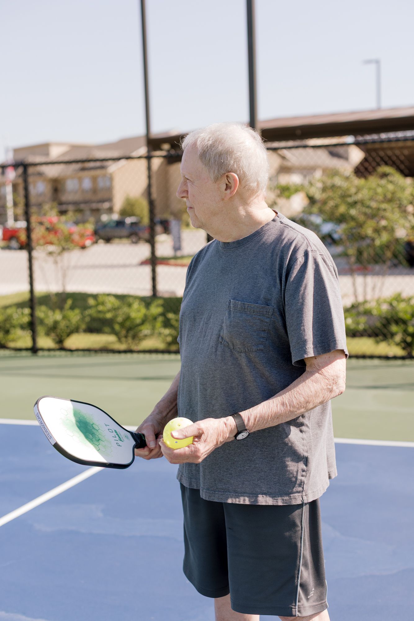 Senior man holding a pickleball paddle and ball on an outdoor blue court on a sunny day.