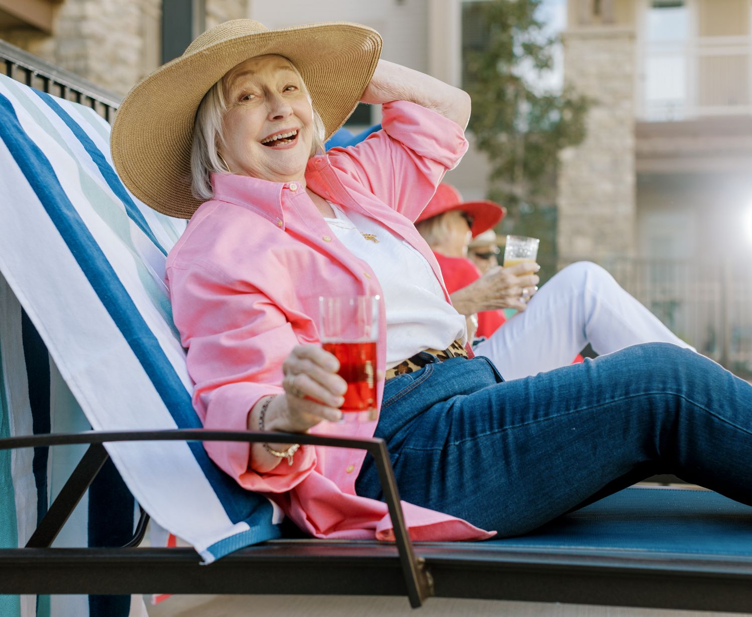 Happy senior woman relaxing on a lounge chair outdoors holding a drink in pink jacket.