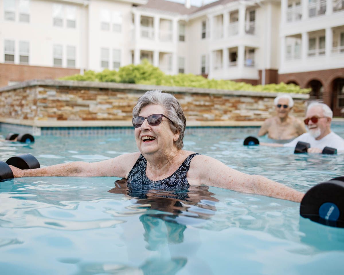 Smiling senior woman doing water aerobics in outdoor pool with other elderly participants using foam dumbbells.