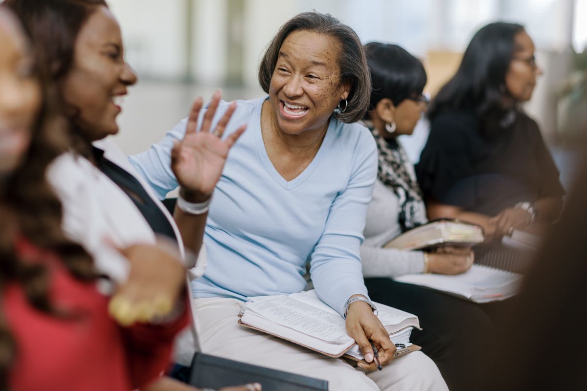 Smiling senior Black woman waving in a classroom setting, engaged in a group learning session with other adults.