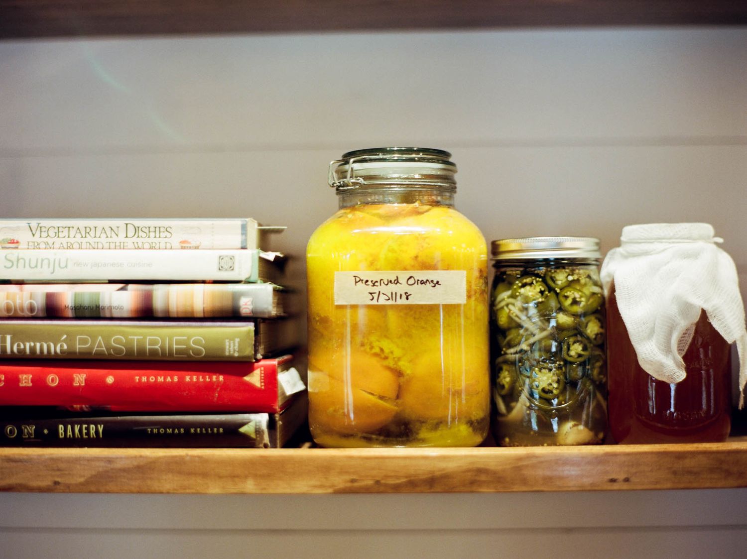 A wooden shelf displaying cookbooks alongside labeled mason jars of preserved pickled orange and vegetables.