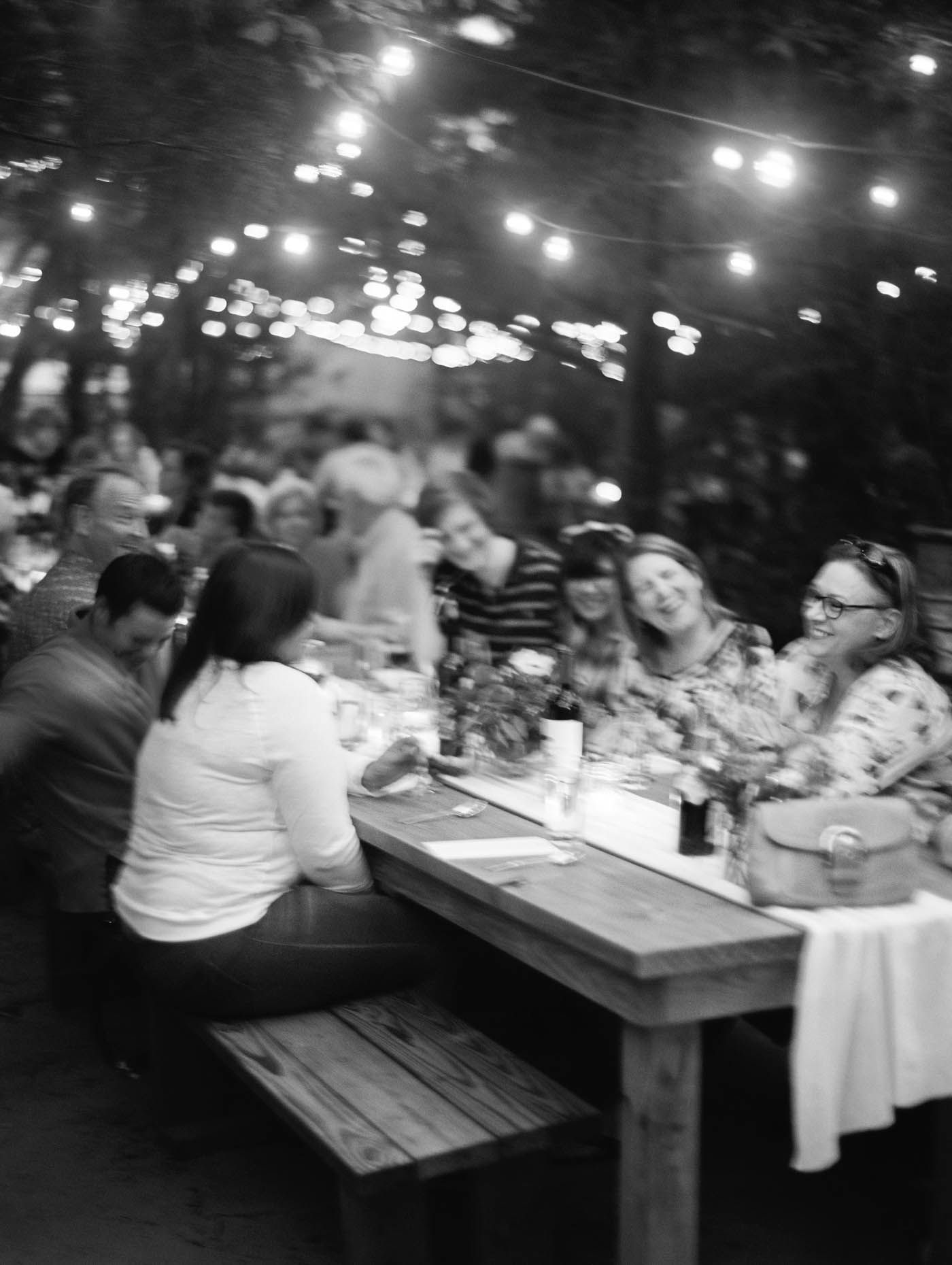 Black and white photo of guests laughing and socializing at an outdoor evening dinner party under glowing string lights.