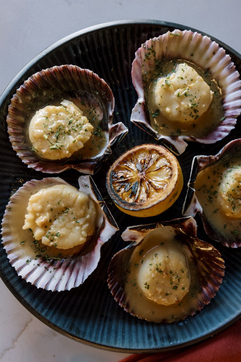 Six baked scallops in shells with herb butter sauce, arranged around a grilled lemon half on a blue plate.