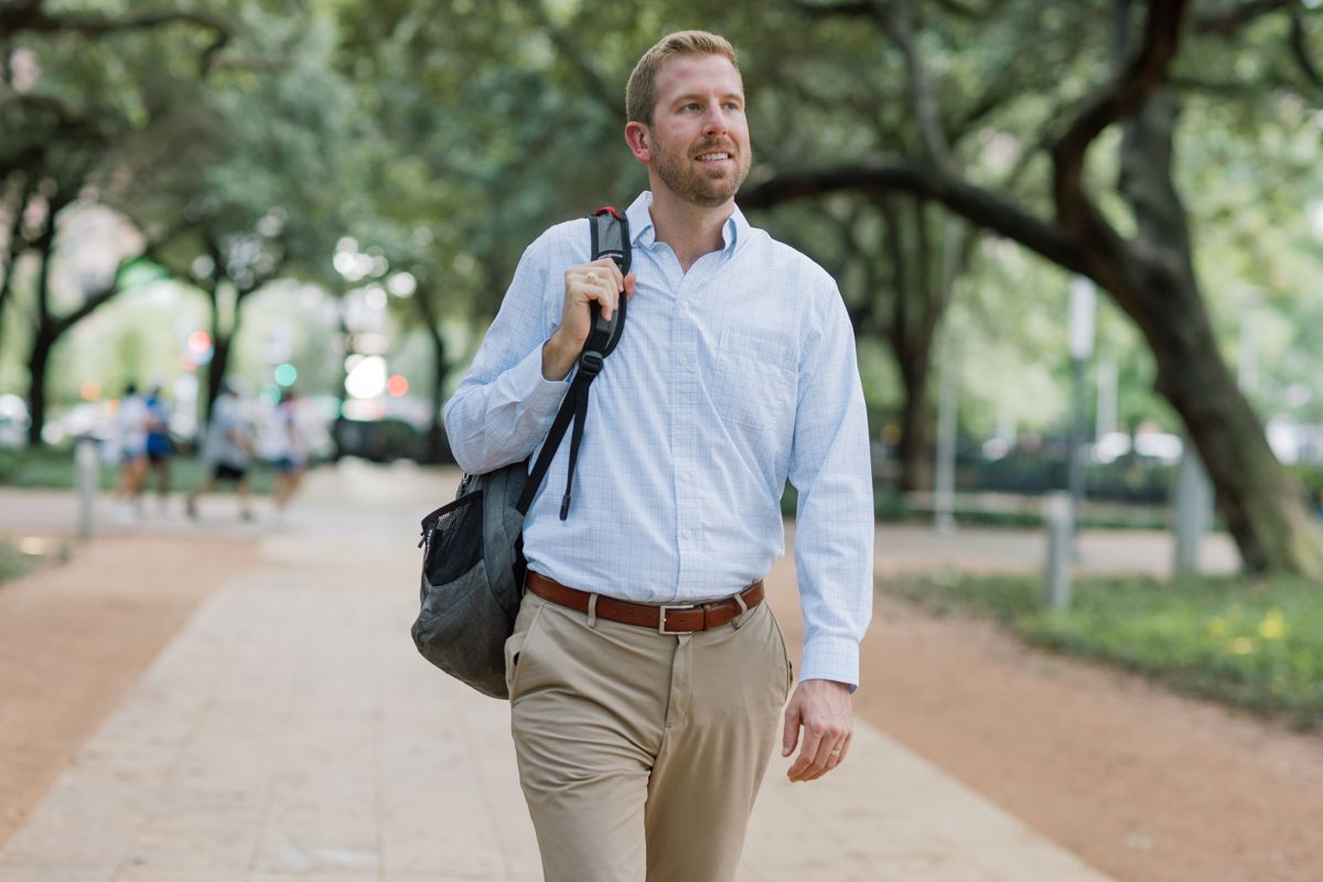 Smiling man with backpack and bag walking along a tree-lined campus pathway.