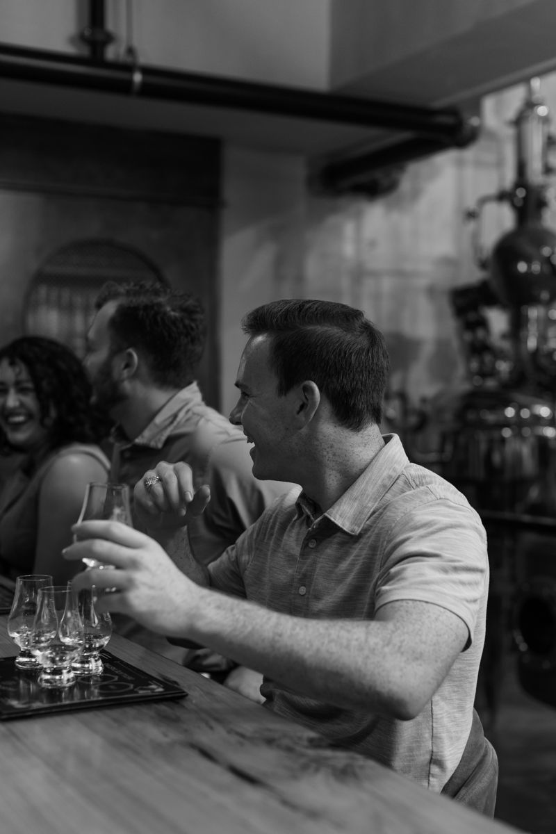 Black and white photo of man examining whiskey flight at distillery bar while friends laugh in background.