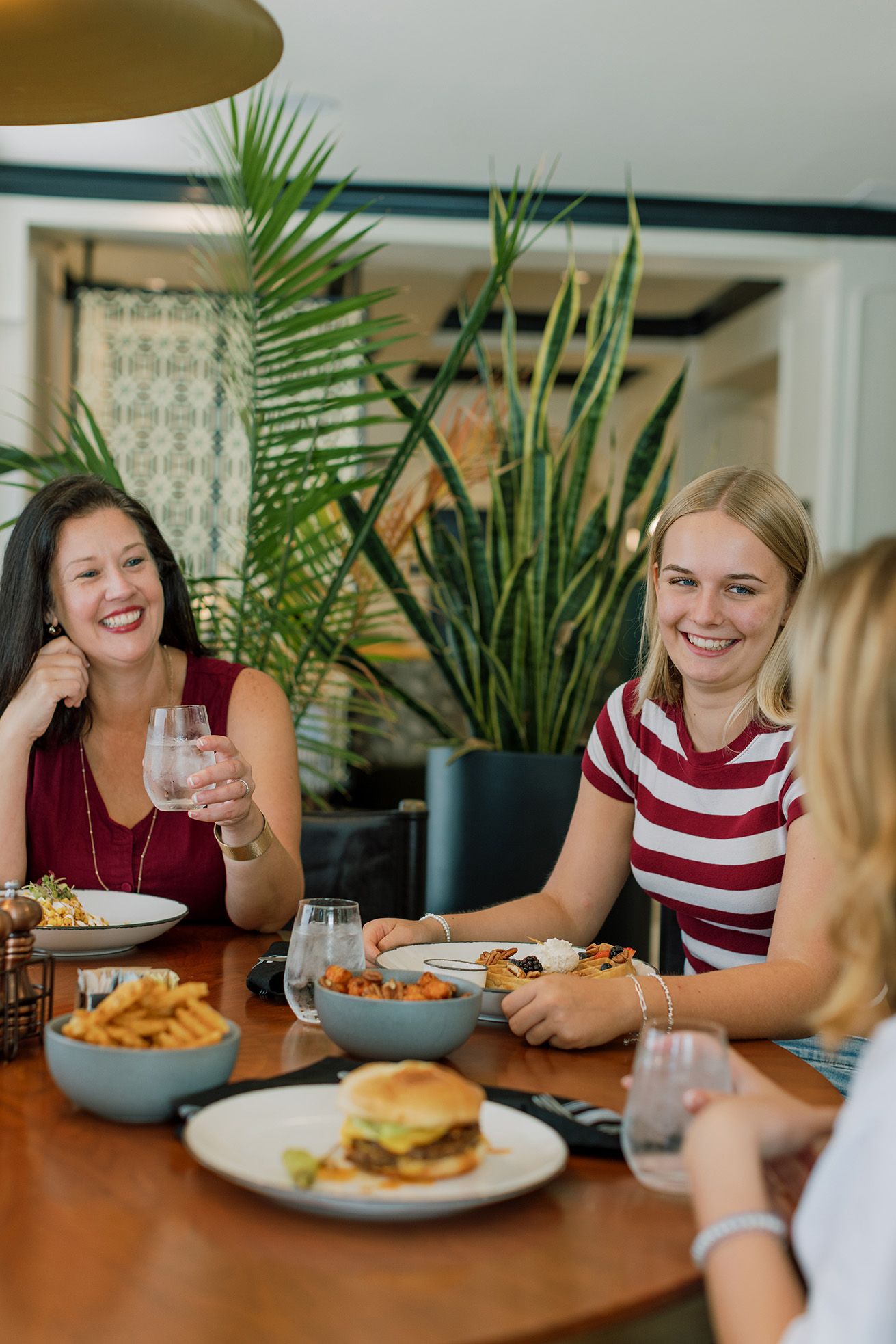Three women laughing and dining together at a restaurant table with burgers, fries, and drinks.