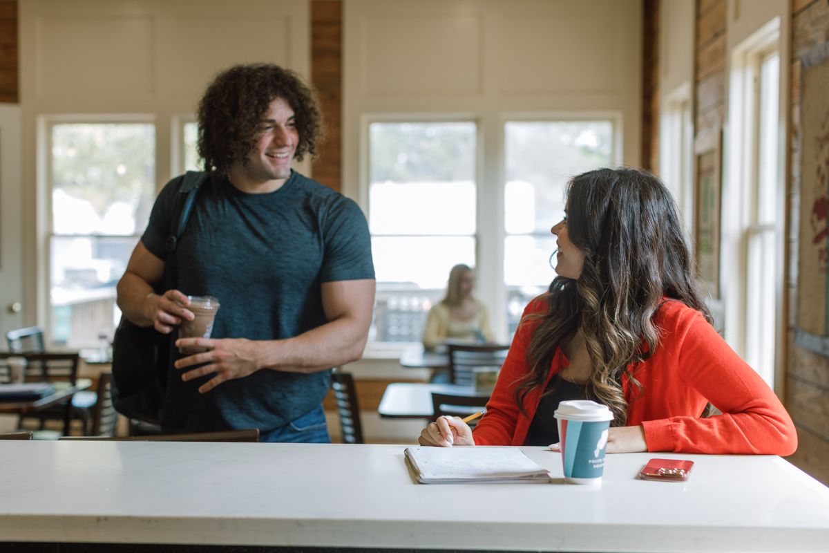 Young man with curly hair chats with a woman in red at a bright cafe table with coffee drinks.