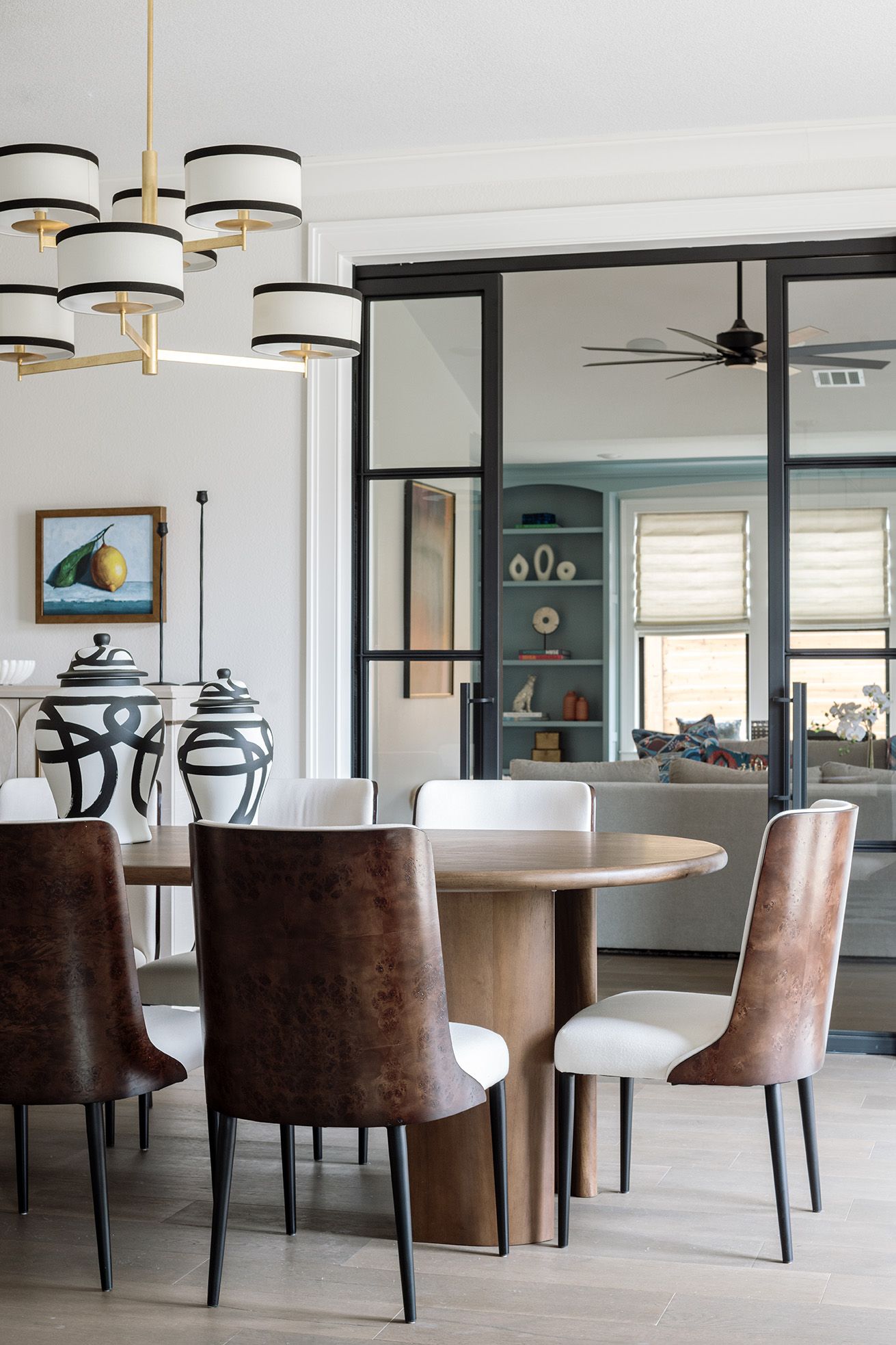Contemporary dining area with round wood table, leather chairs, striped chandelier, and steel-framed glass partition.
