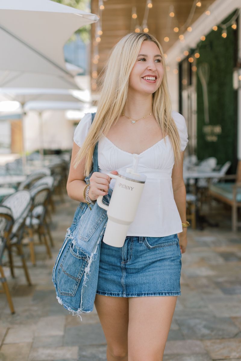 Smiling blonde woman in white top and denim skirt carries a large tumbler and denim bag outdoors.