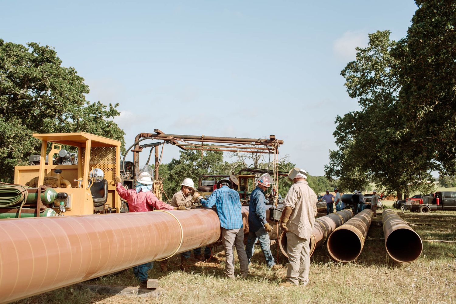 Workers assembling large industrial pipes outdoors using heavy machinery on a rural construction site.